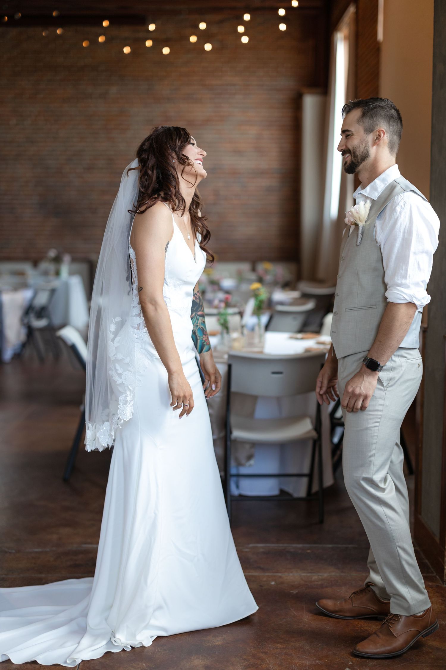 Candid wedding moment in rustic venue with white lace gown and gray suit against brick wall backdrop.