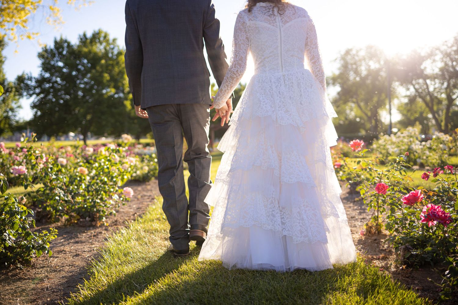 Newlyweds walking hand in hand through garden path at sunset.