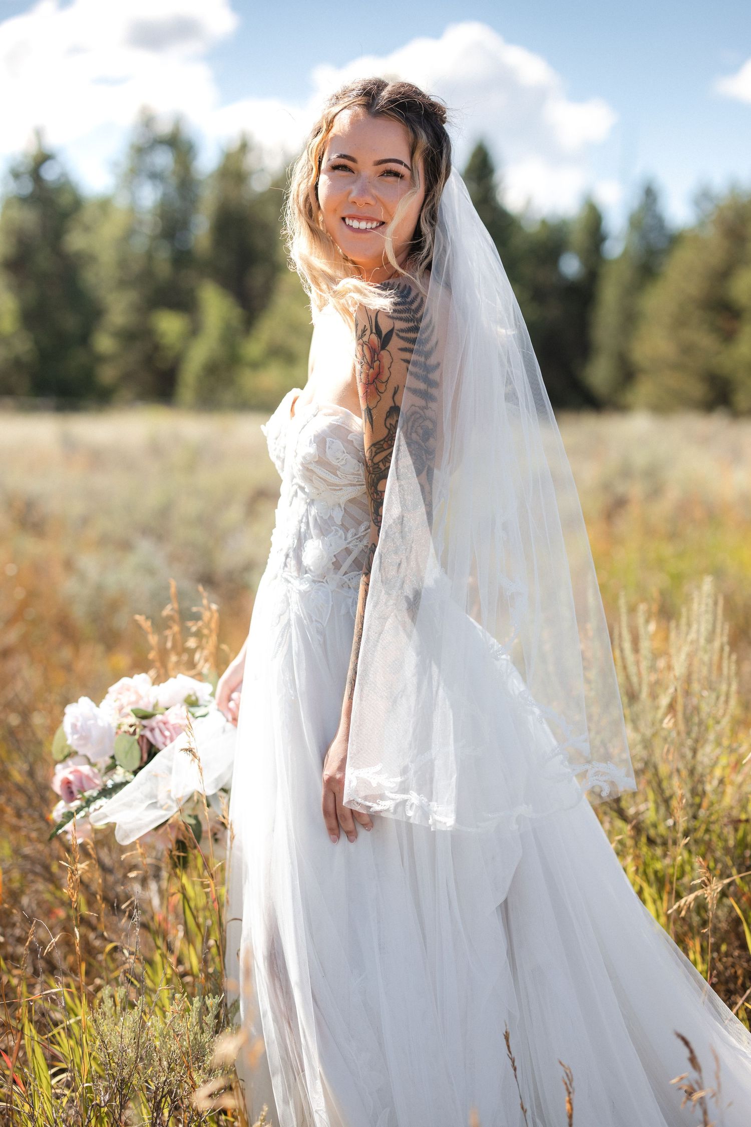 A bride with a delicate veil poses in a sunlit meadow holding a pink and white bouquet.