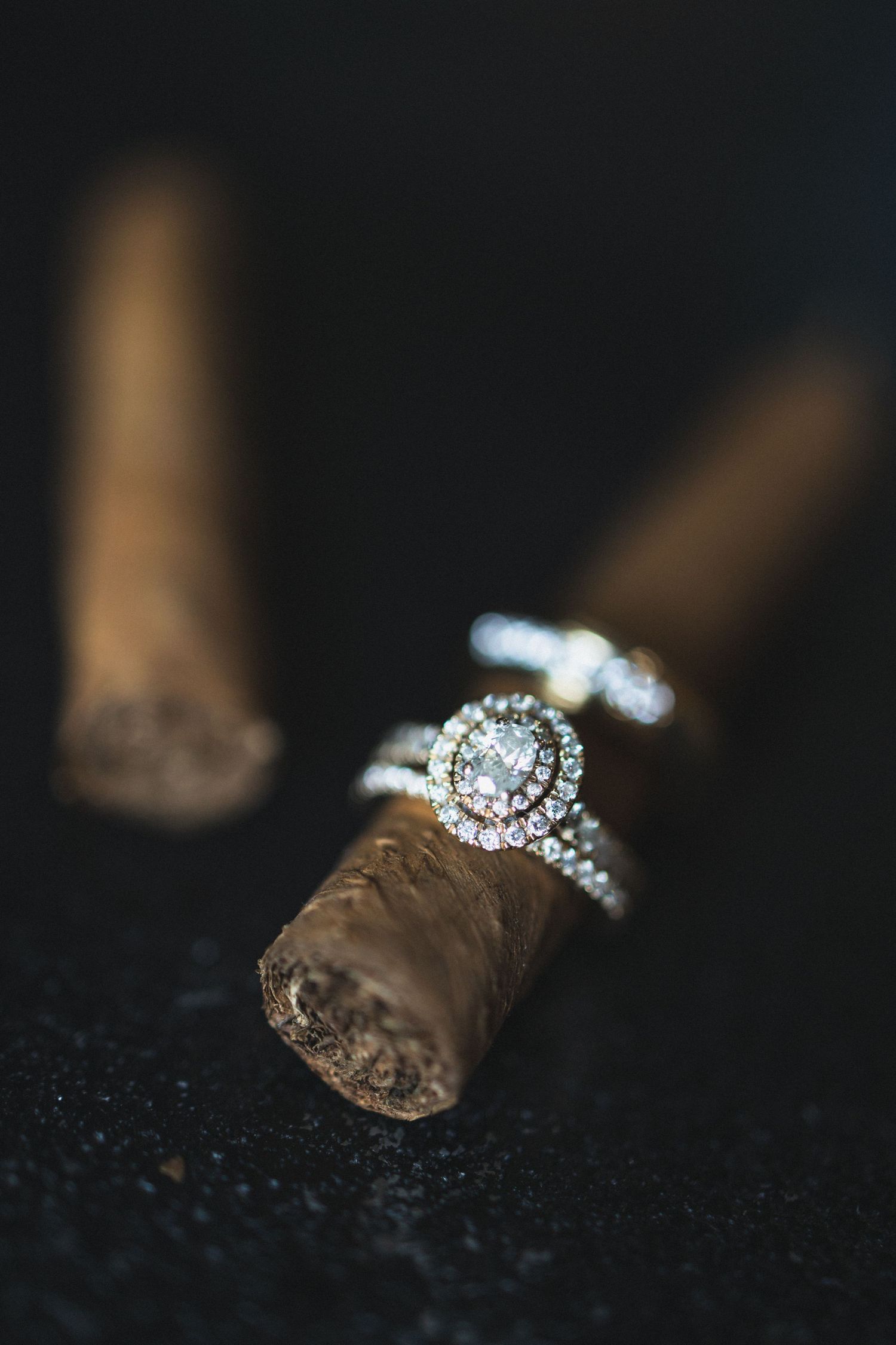 Elegant diamond engagement ring perched on brown cigar against dark background.