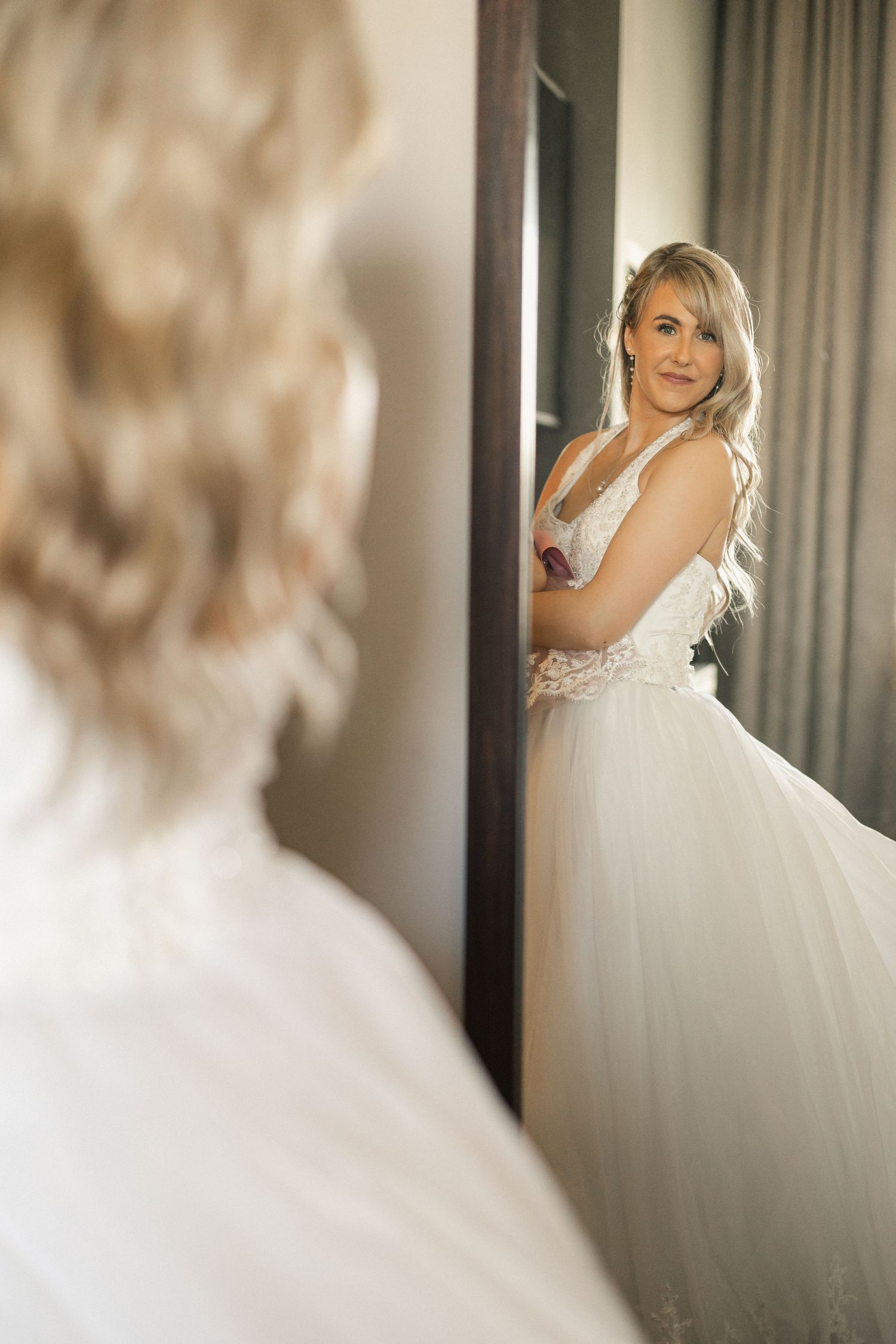Bride in white lace gown admiring her reflection in a full-length mirror.
