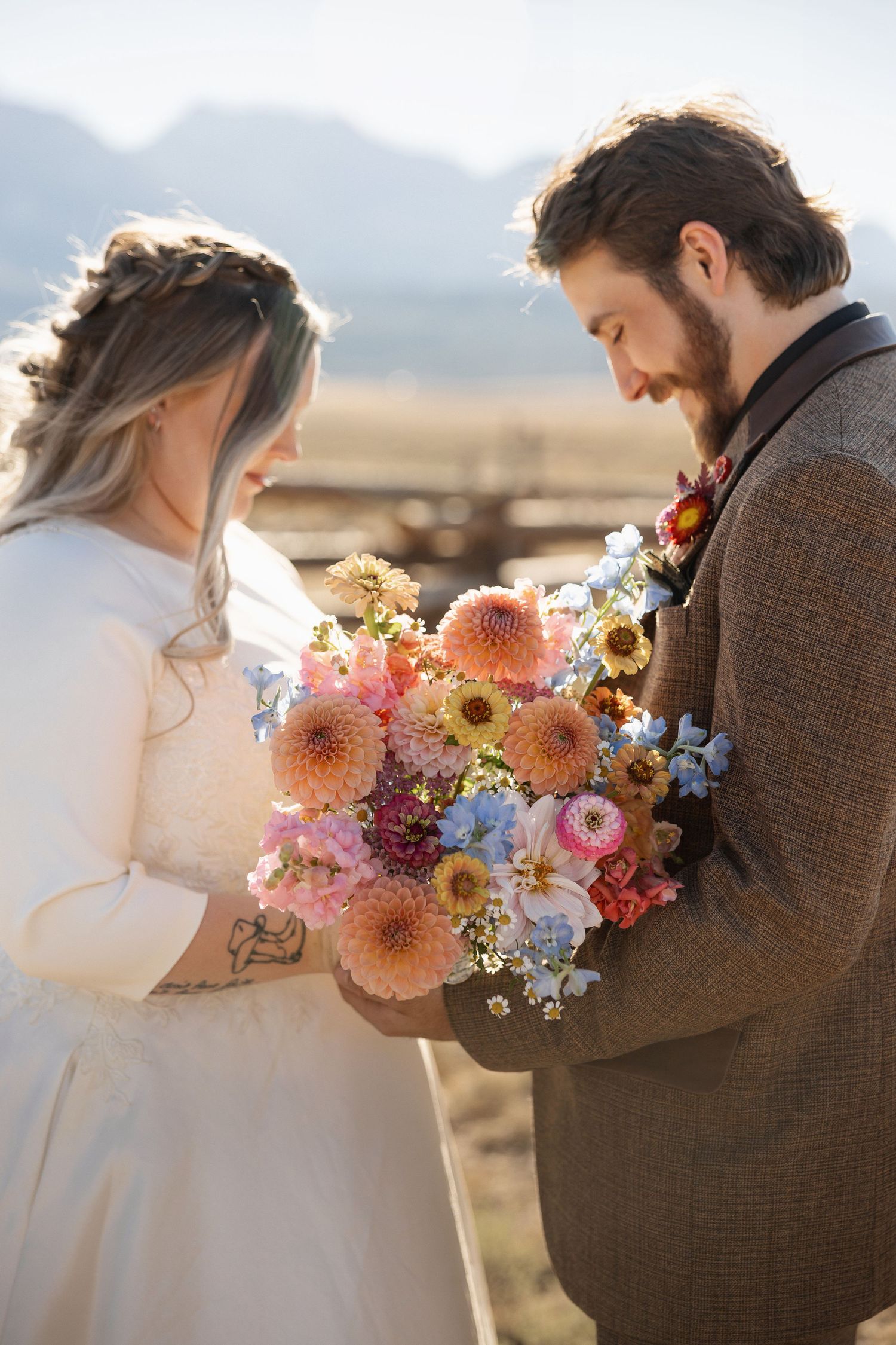 A couple shares an intimate moment with a stunning bouquet of pink dahlias and blue flowers against a mountain backdrop.