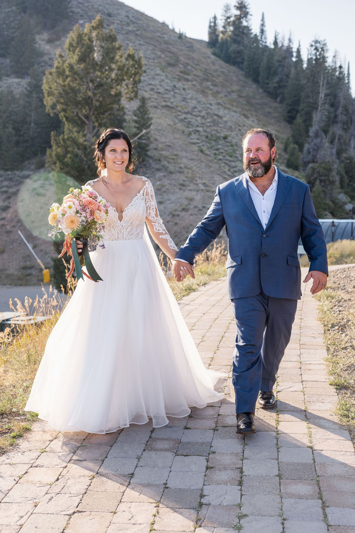Couple walking together on a stone pathway with mountains and pine trees in the background.