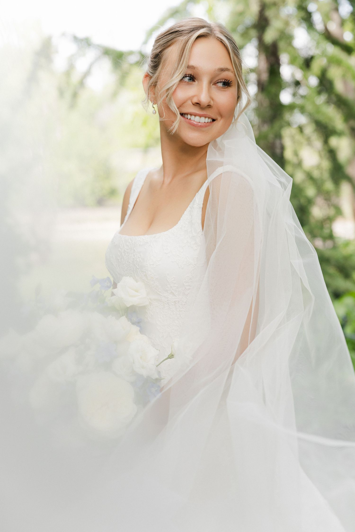 Smiling bride in white lace gown and veil holding white floral bouquet outdoors.