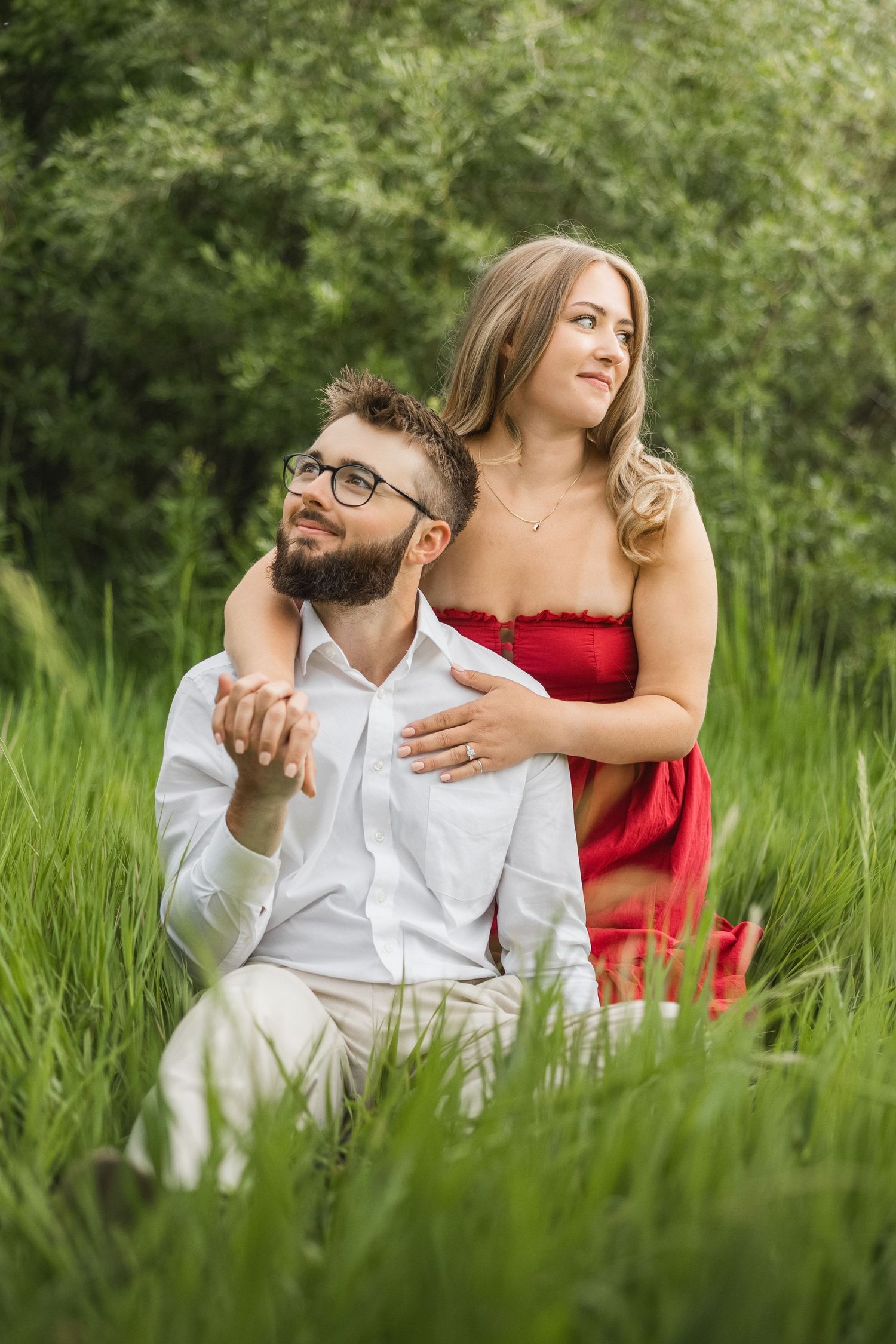 Couple sitting in tall green grass, woman in red dress embracing man in white shirt wearing glasses outdoors.