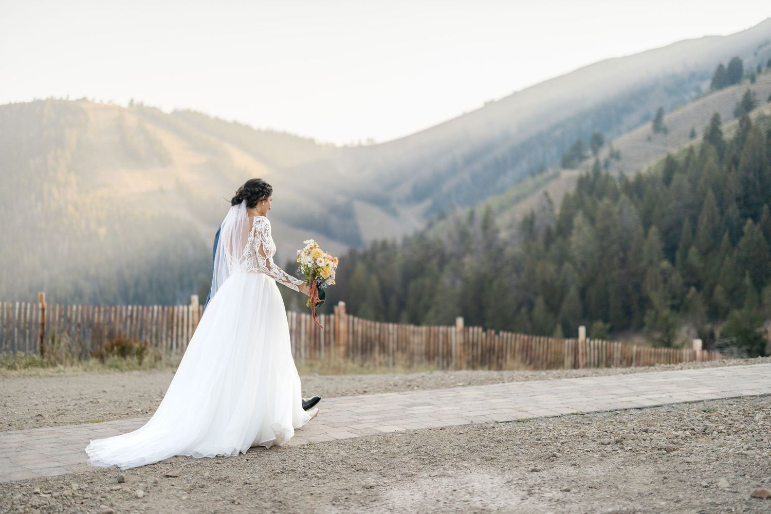 A bride in a flowing white gown holds a bouquet while standing on a mountain road at sunset with hills in the background.