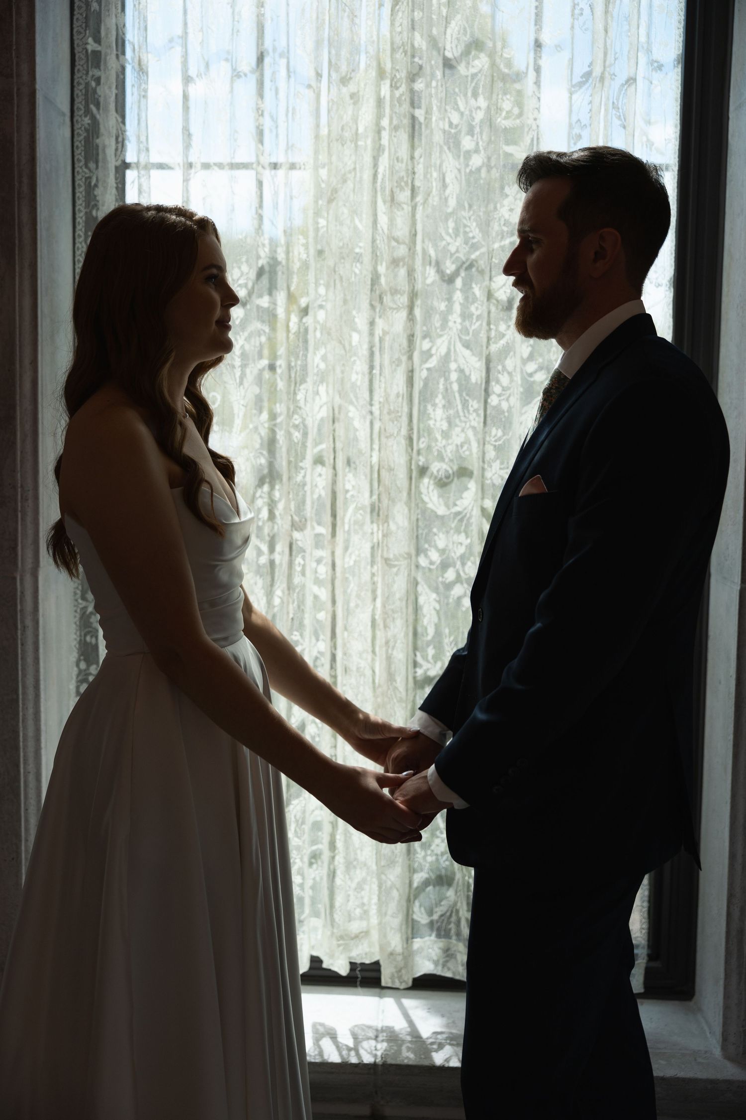 Silhouetted wedding couple shares tender moments by window with lace curtains.