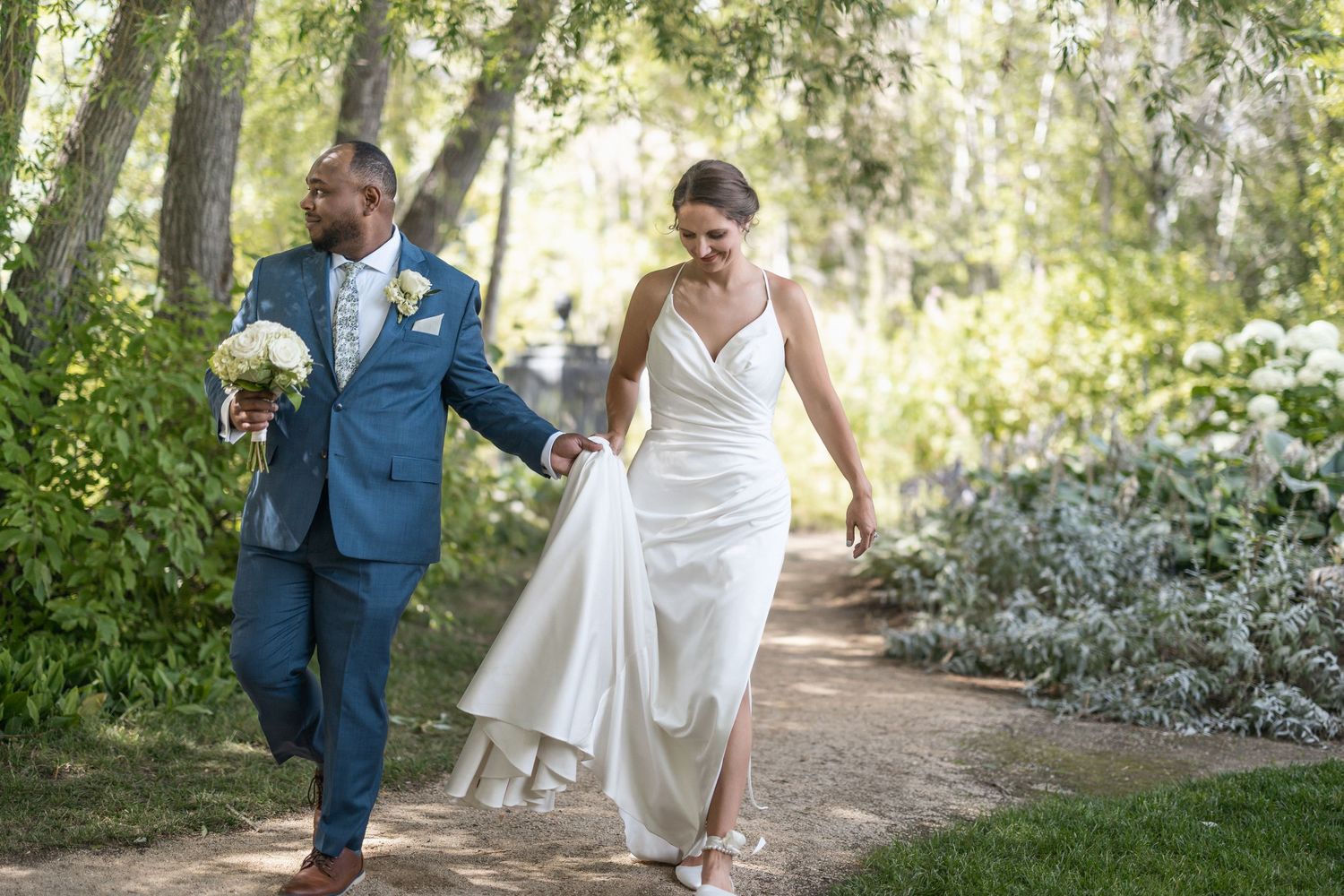 A couple shares a romantic moment on a garden path surrounded by lush greenery and trees.