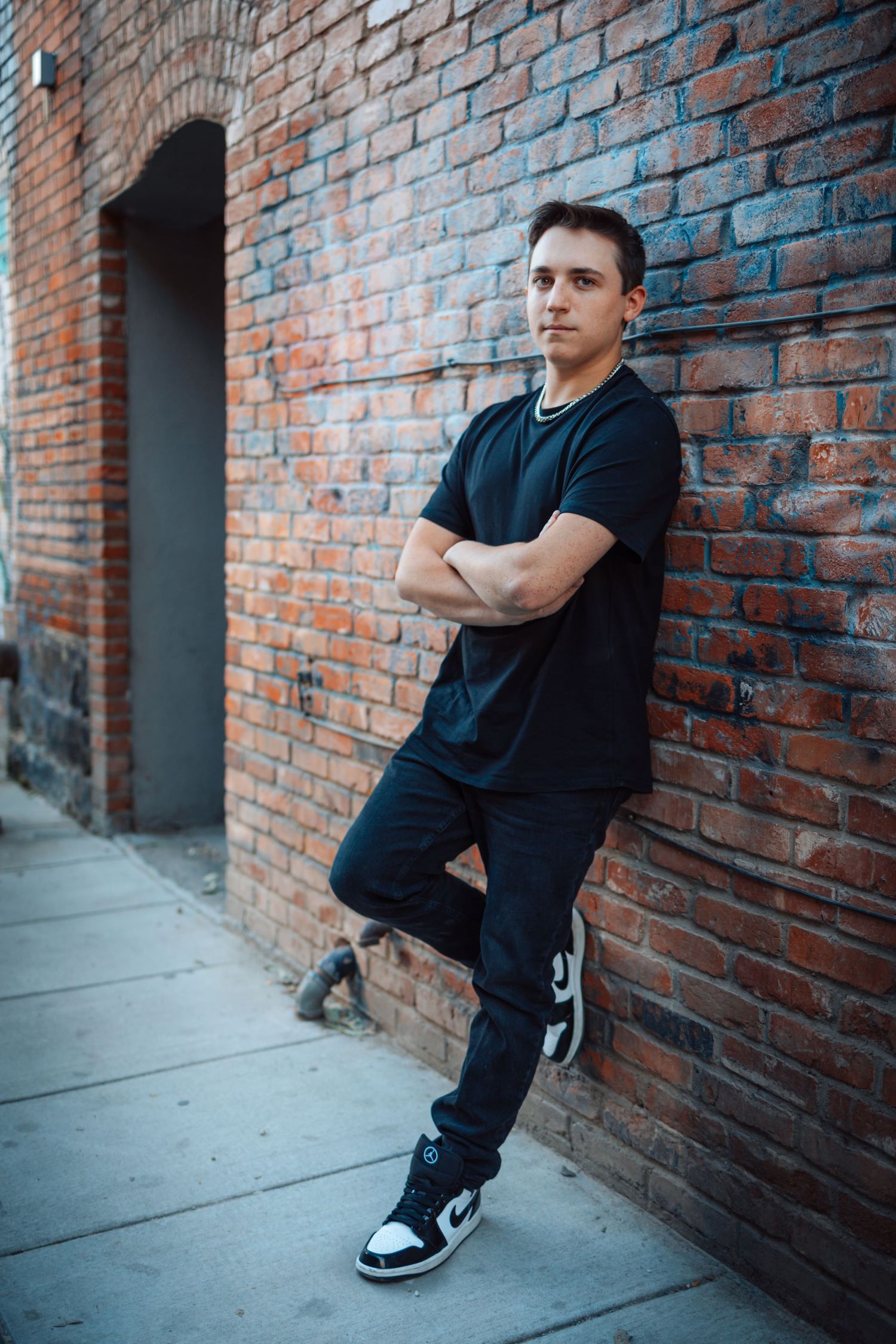 Person in all black attire posing against a brick wall in an urban setting, shown in multiple angles.