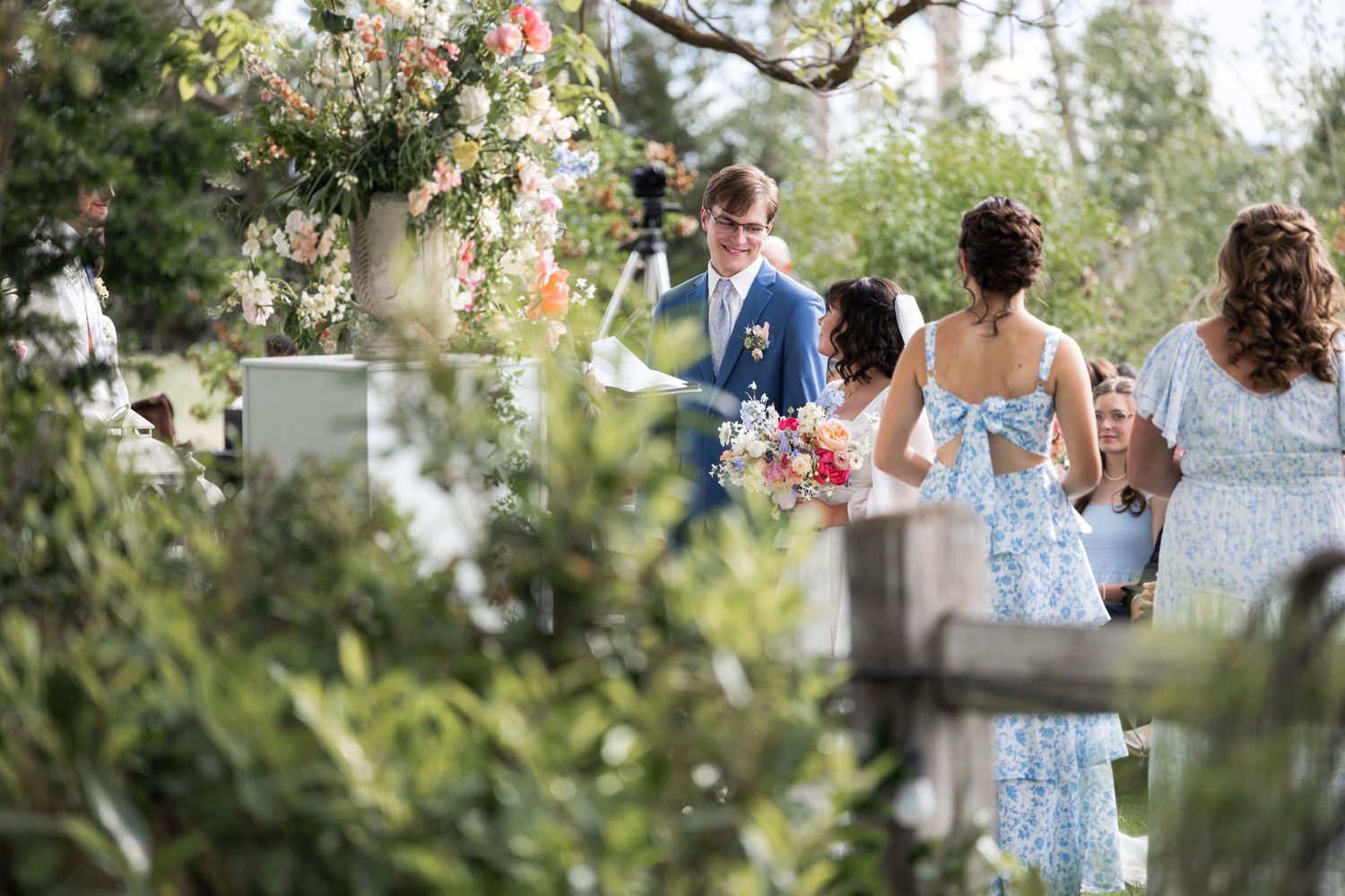 Wedding party members stand among floral arrangements during outdoor garden ceremony.