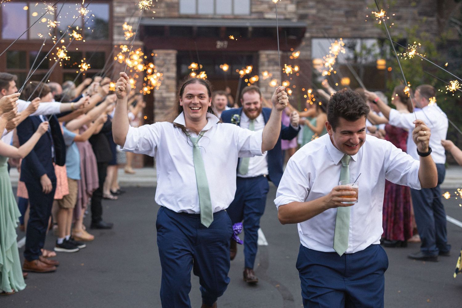 Couple running through sparkler exit tunnel while guests cheer at outdoor wedding celebration.