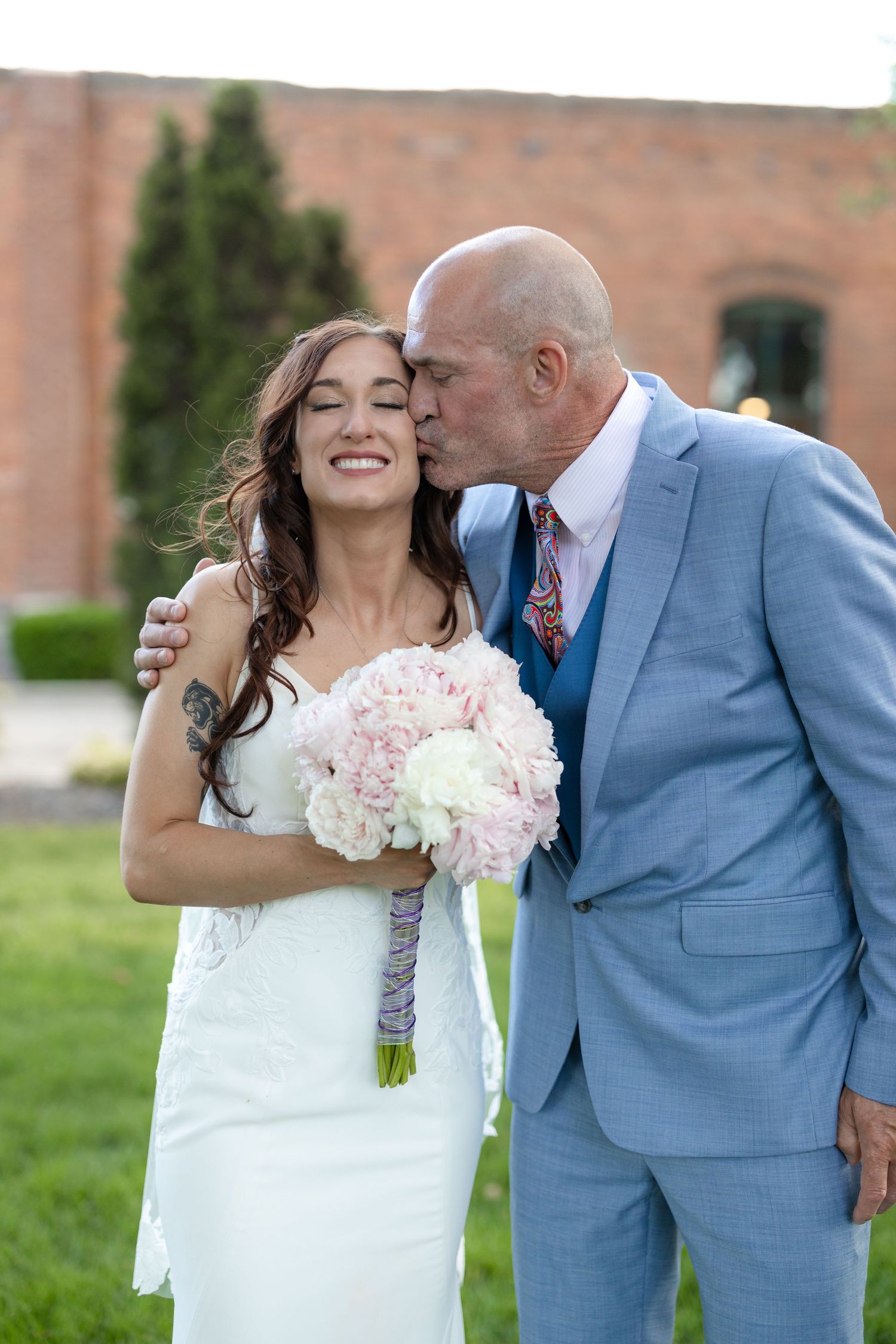 Wedding photo showing tender moment with soft pink bouquet and blue suit against brick backdrop.