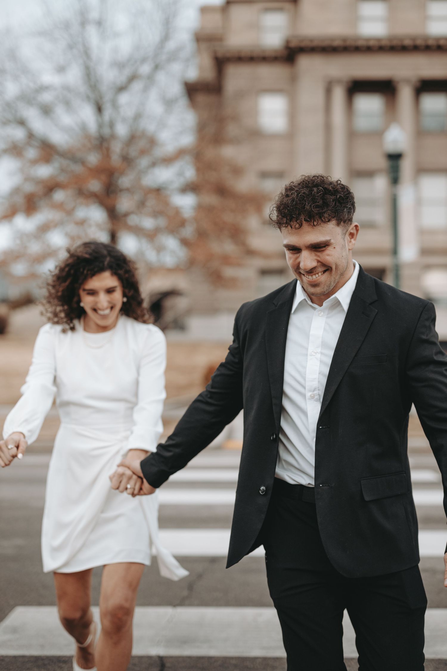 A joyful couple in formal attire share a laugh while walking across a city crosswalk.