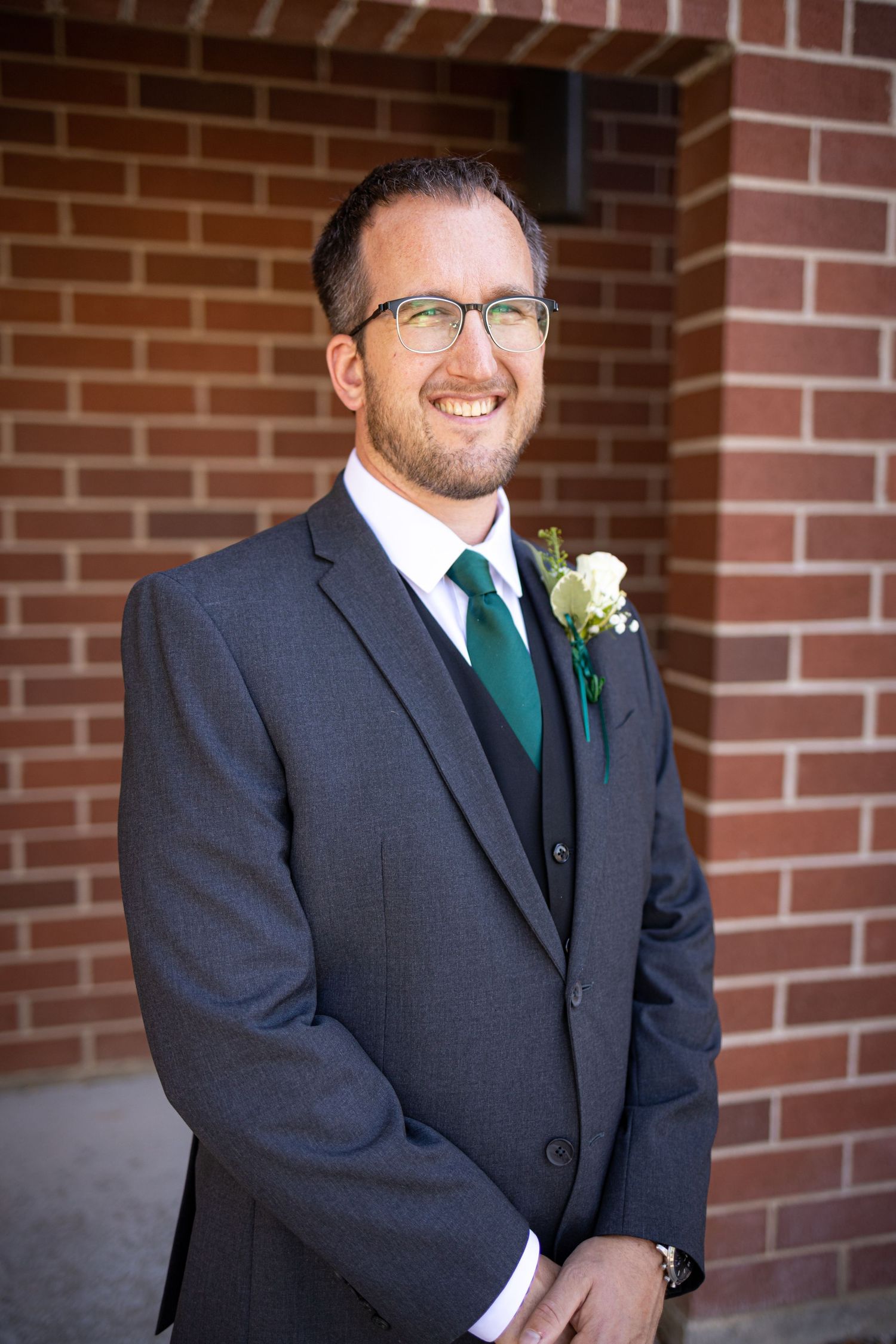 Man in dark suit with emerald tie and white boutonniere poses against brick wall.