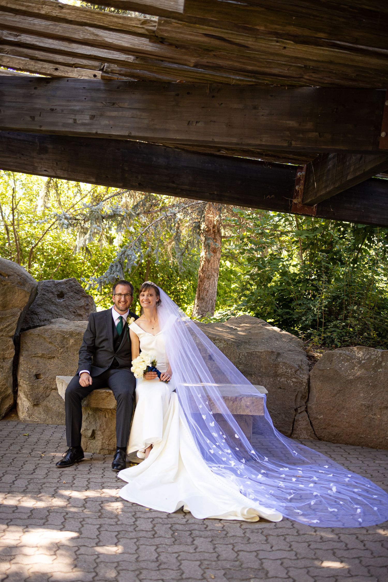 Wedding couple sits on stone bench under rustic wooden pergola with flowing veil.