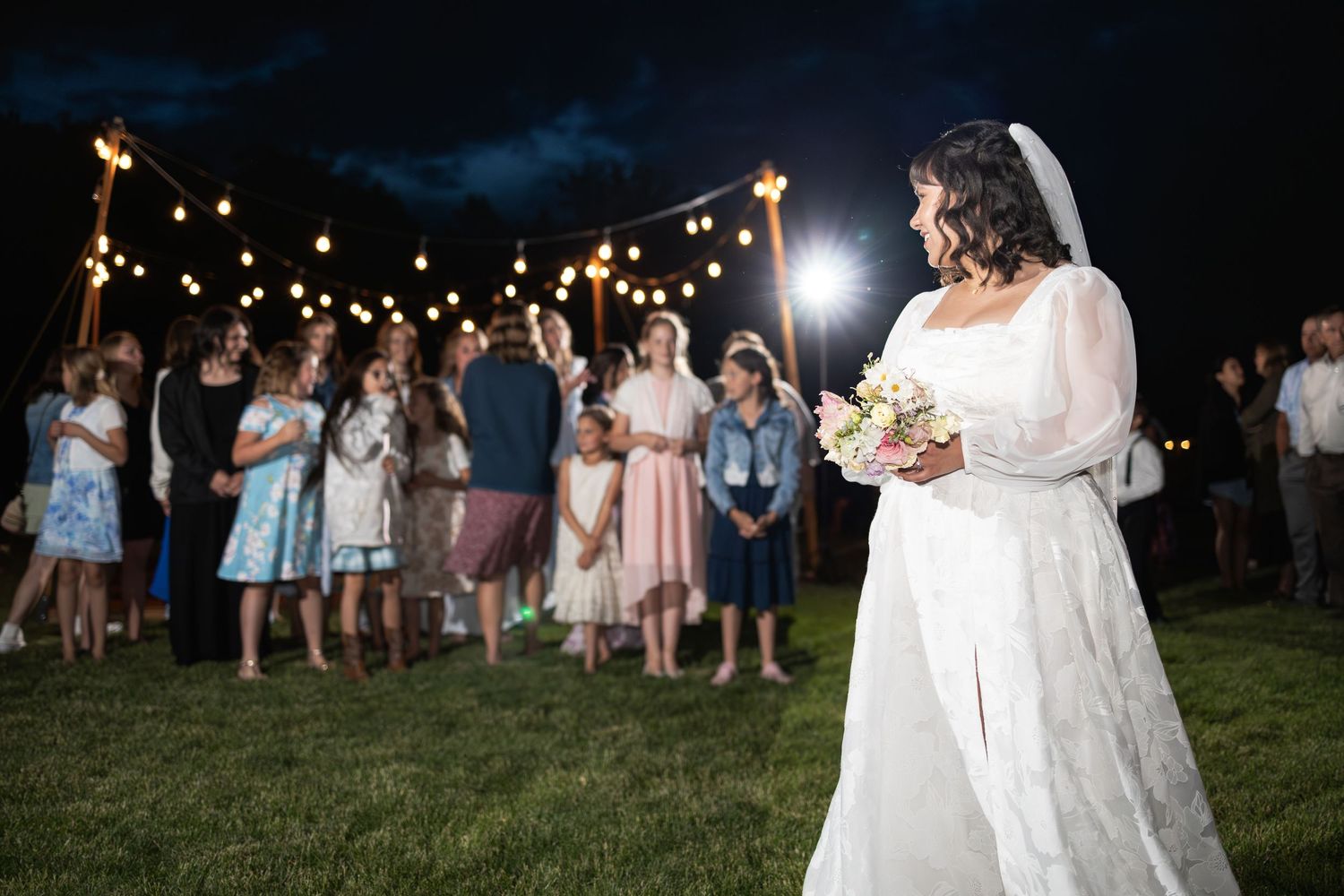 Wedding guests gather beneath string lights at dusk for outdoor festivities.