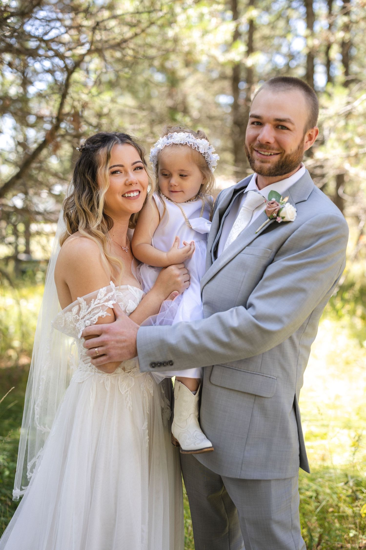 A joyful wedding portrait of a couple and their young child in a sunlit forest.