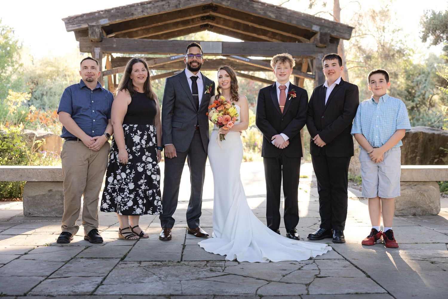 Wedding party poses together under rustic wooden gazebo during outdoor ceremony.