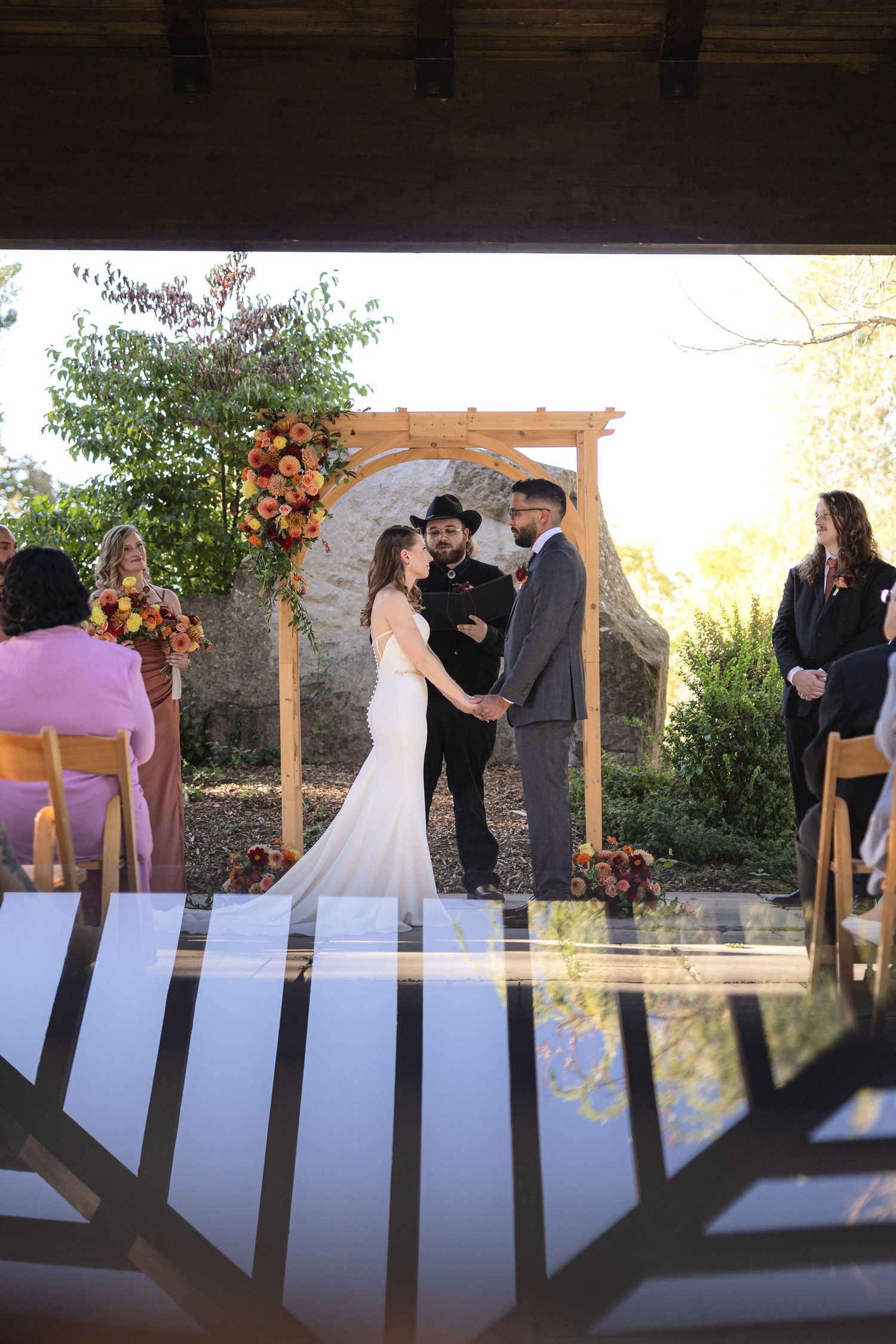 Wedding ceremony under wooden arbor decorated with orange and pink flowers.