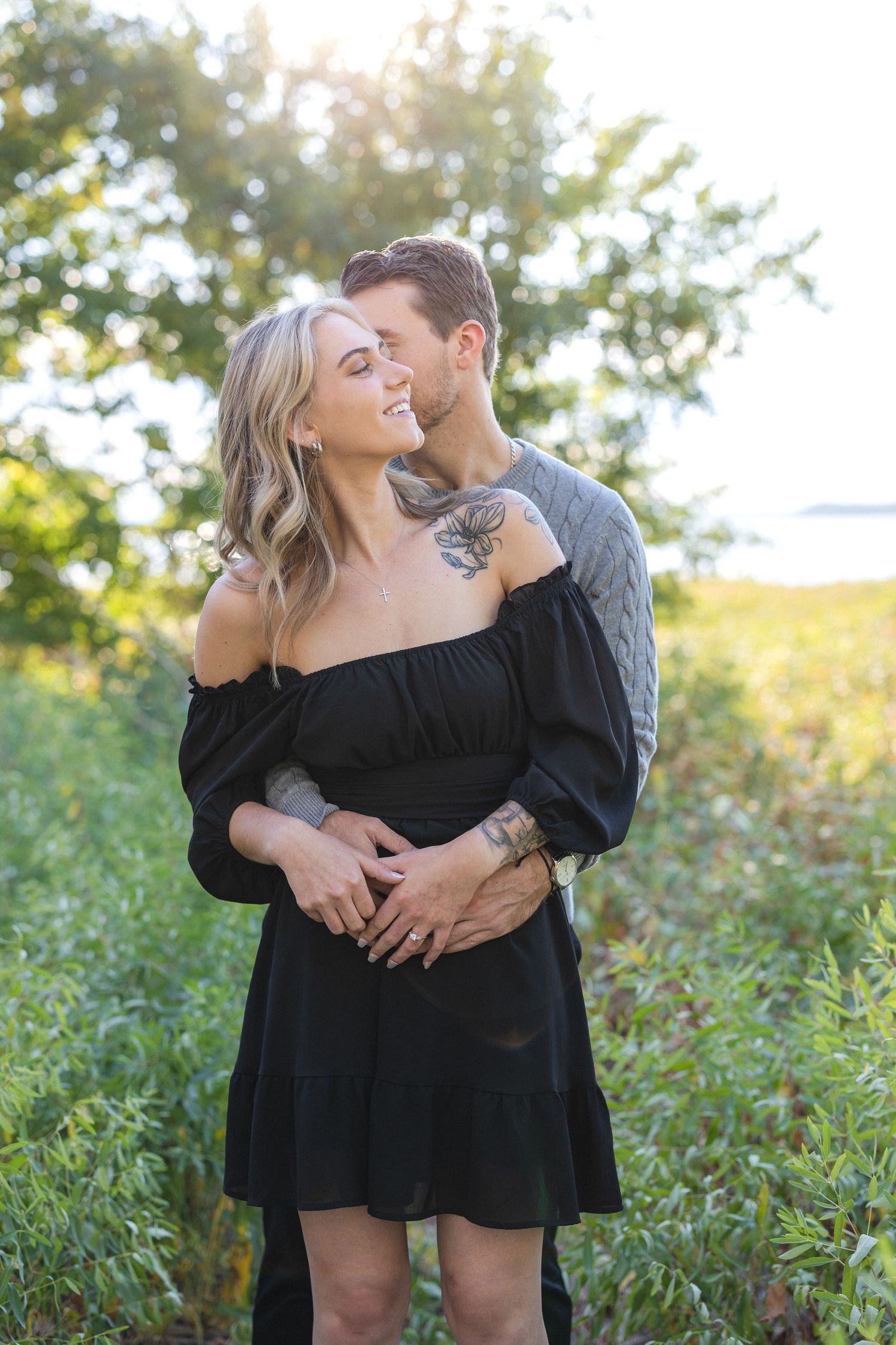 Couple embracing in black dress and grey sweater against natural outdoor setting.