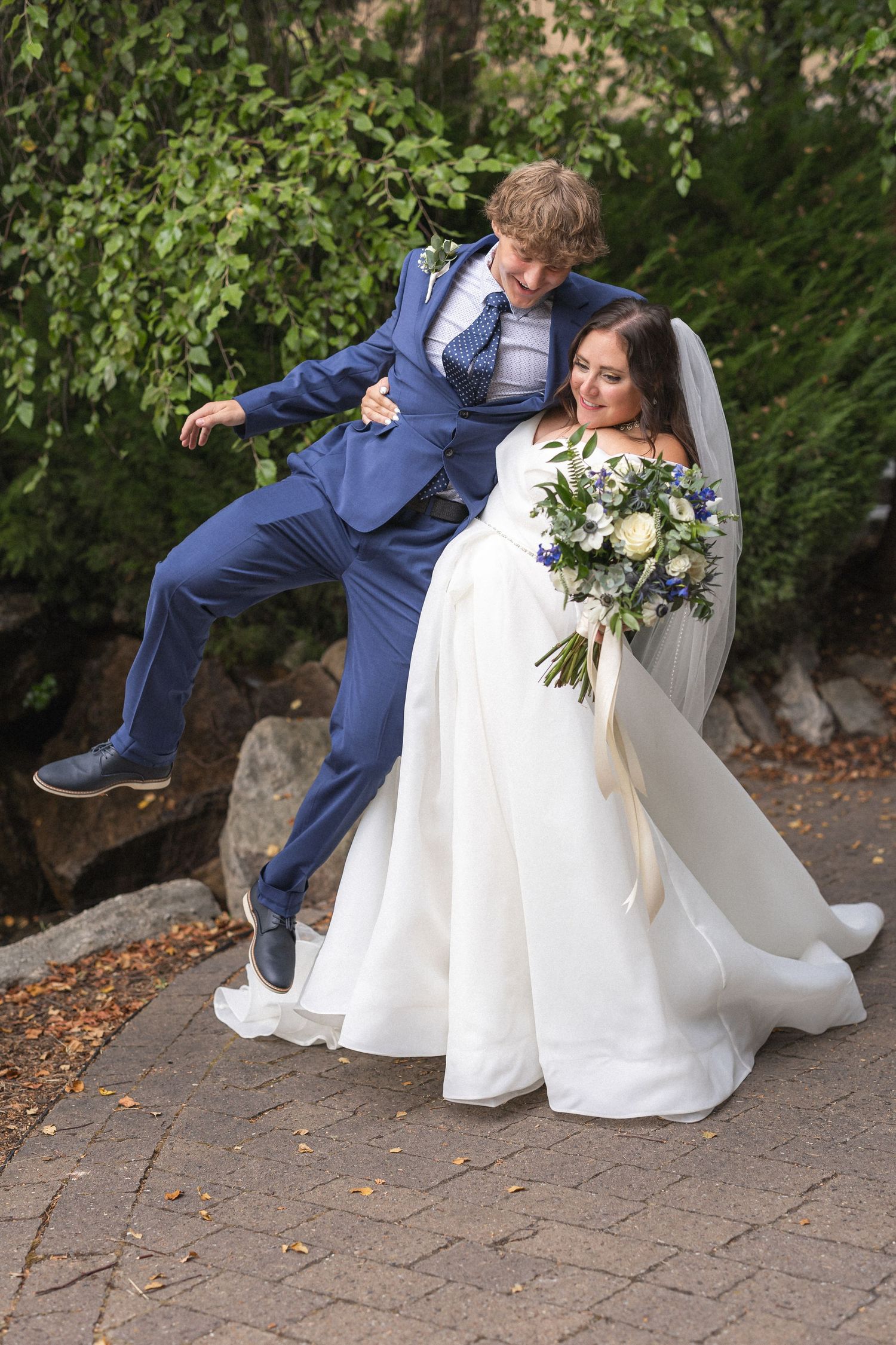 Playful wedding photo with groom jumping while bride holds bouquet on brick pathway.