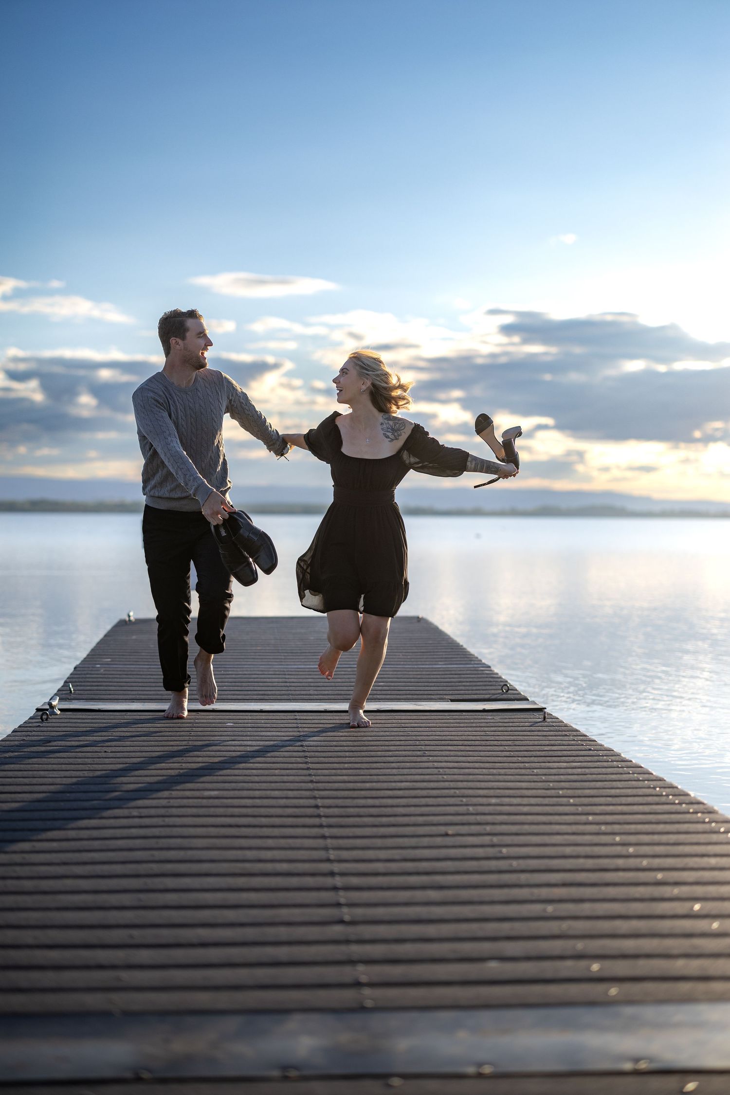 A couple dances barefoot on a wooden pier against a serene waterfront backdrop.