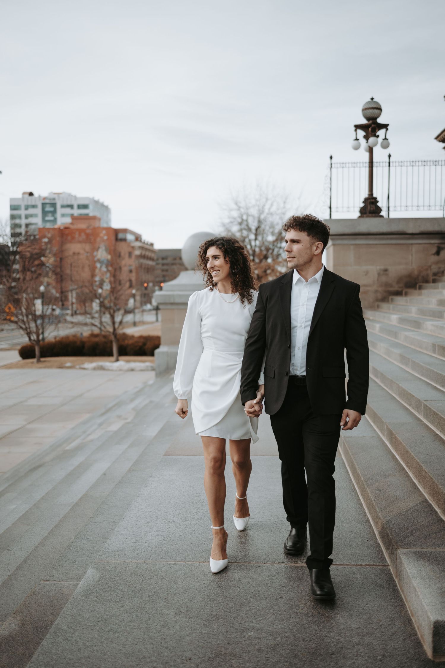 A couple walks down urban steps together in formal attire against a city backdrop.