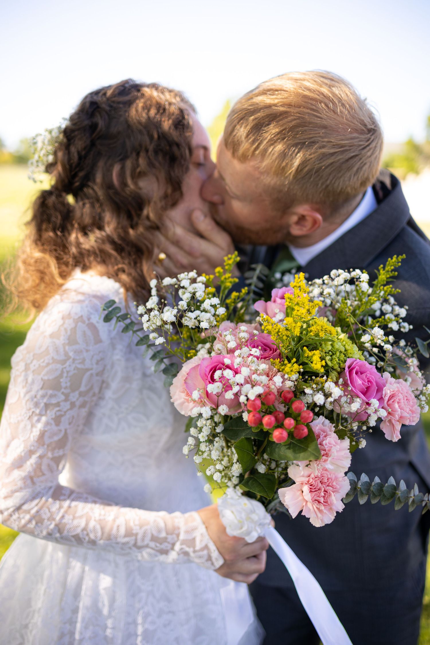 An intimate wedding photo showing a romantic kiss while holding a vibrant bouquet of pink and yellow flowers.