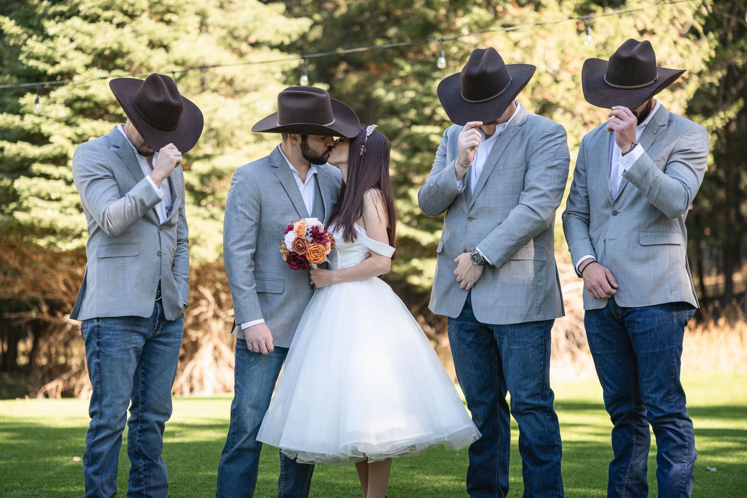 Groomsmen in light blue suits and cowboy hats share a lighthearted moment with the bride in a tea-length dress.