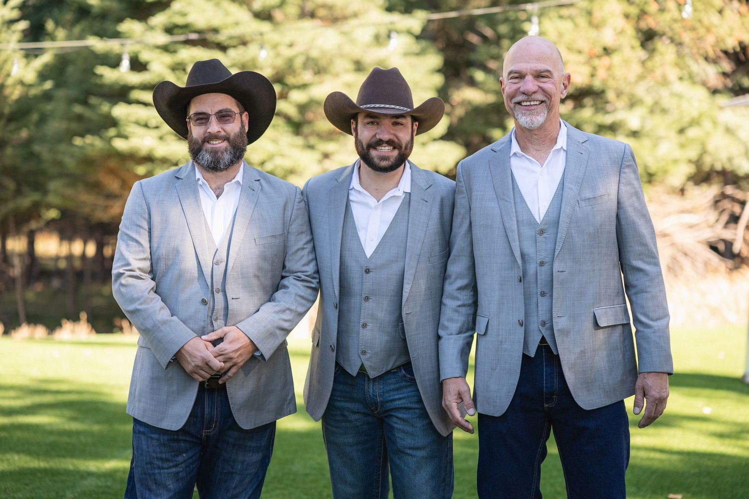 Three men in matching gray suits and cowboy hats pose at outdoor wedding.