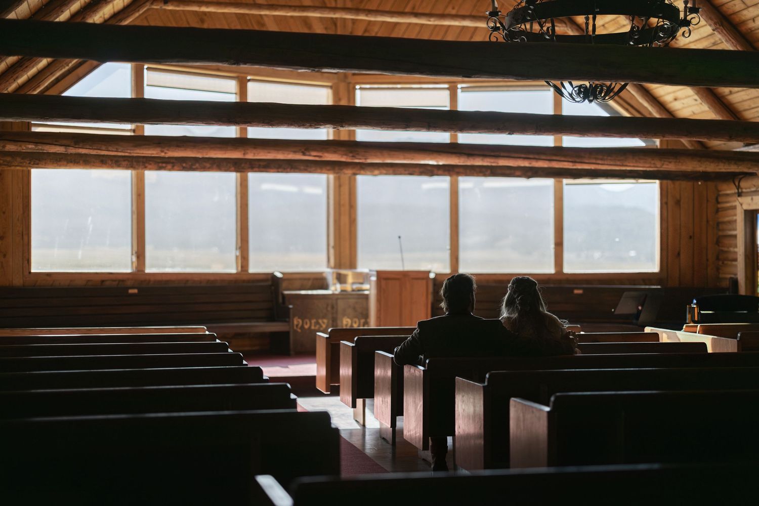 Interior view of rustic wooden chapel with rows of empty pews and large windows letting in natural light.