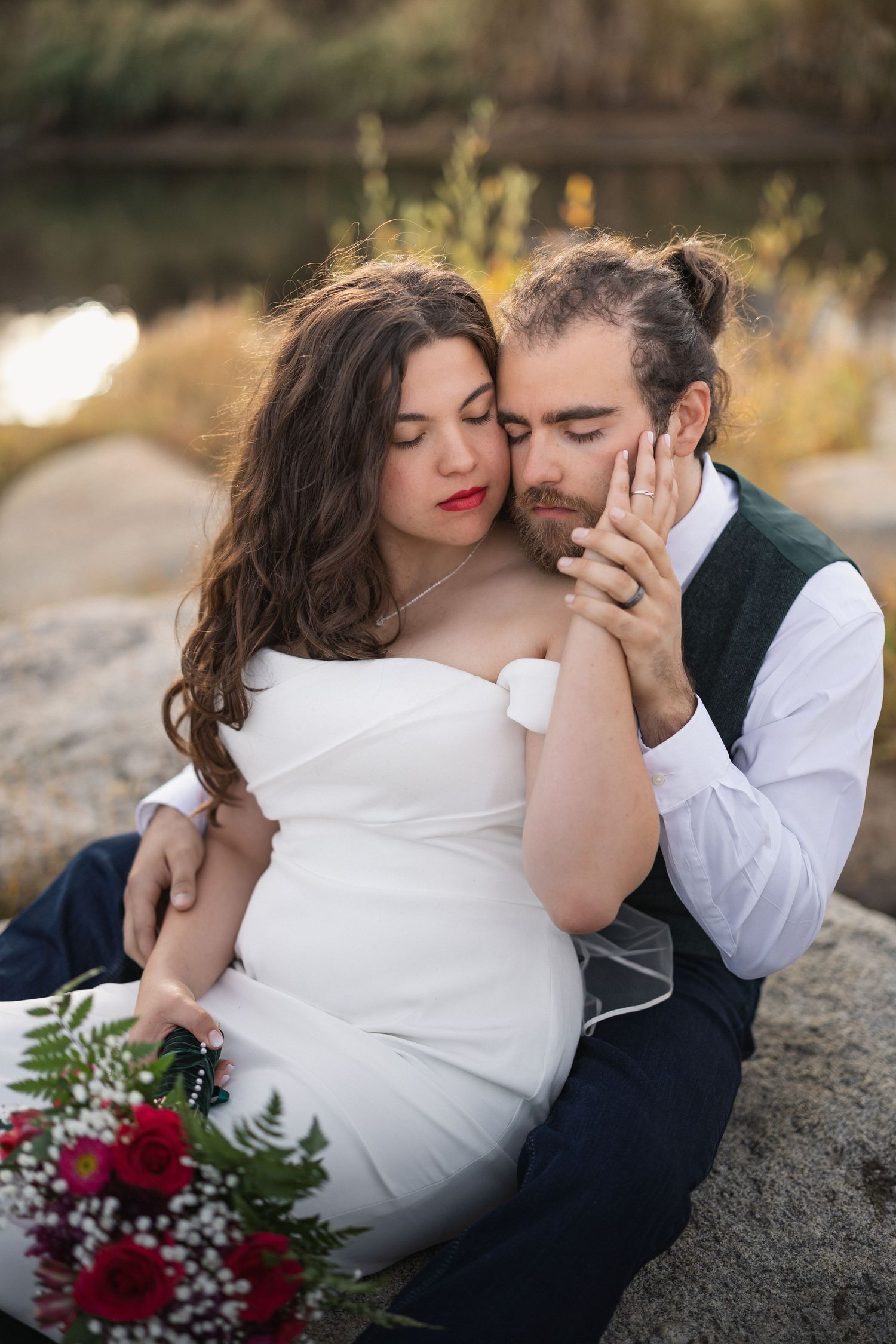 Intimate moment captured on rocks by water with red roses and autumn scenery.