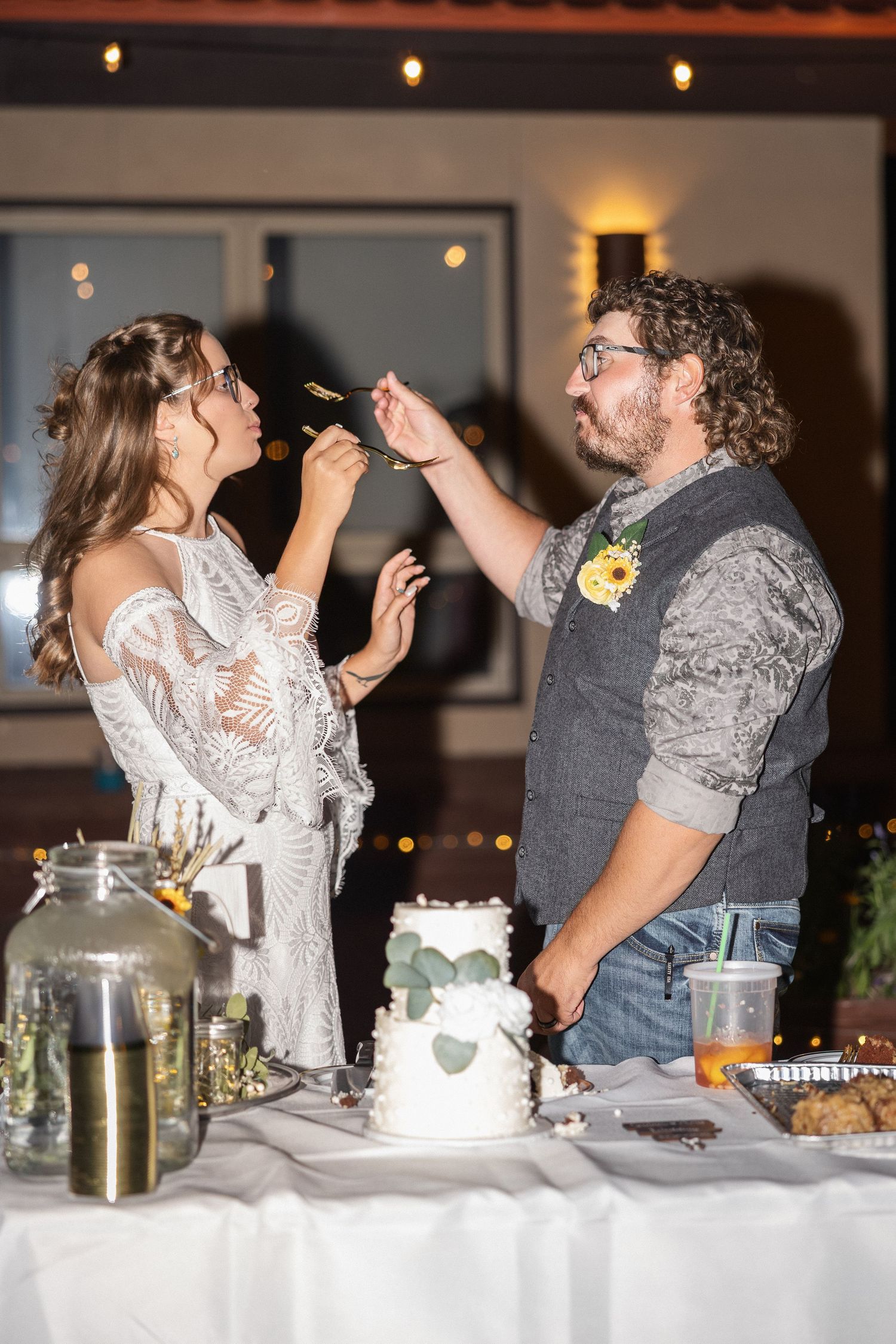 A couple shares cake during their wedding reception indoors under warm lighting.