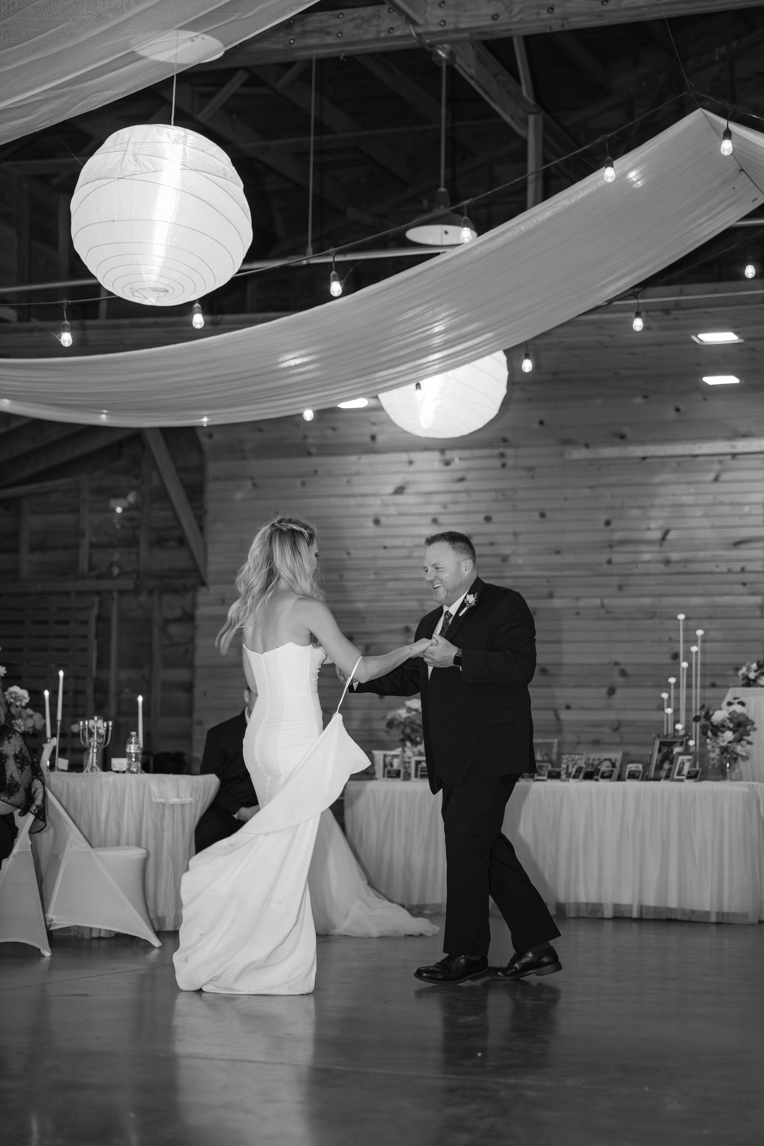 A couple dances in a rustic reception venue decorated with paper lanterns and fabric draping.