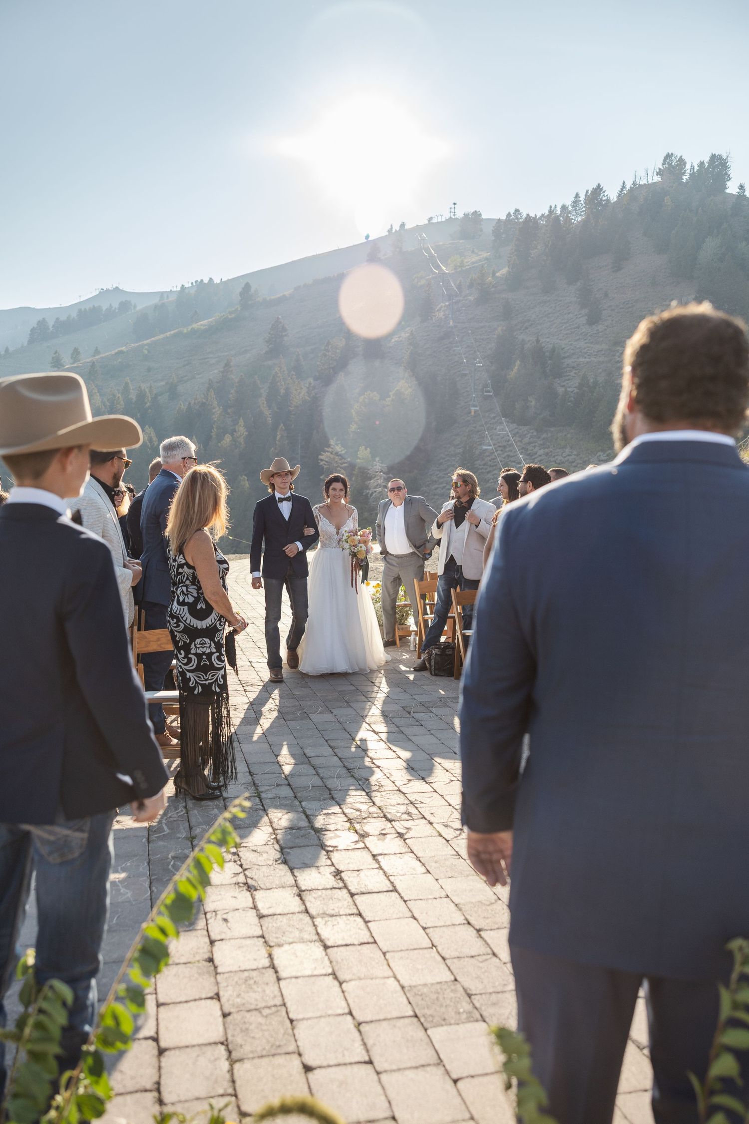 Wedding processional on stone patio with mountains and forest in background during golden hour.