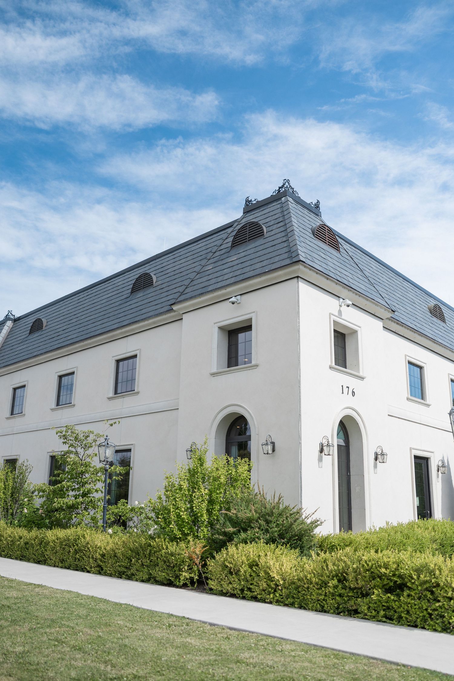A white French-style mansion with gray roof and manicured landscaping against blue sky.
