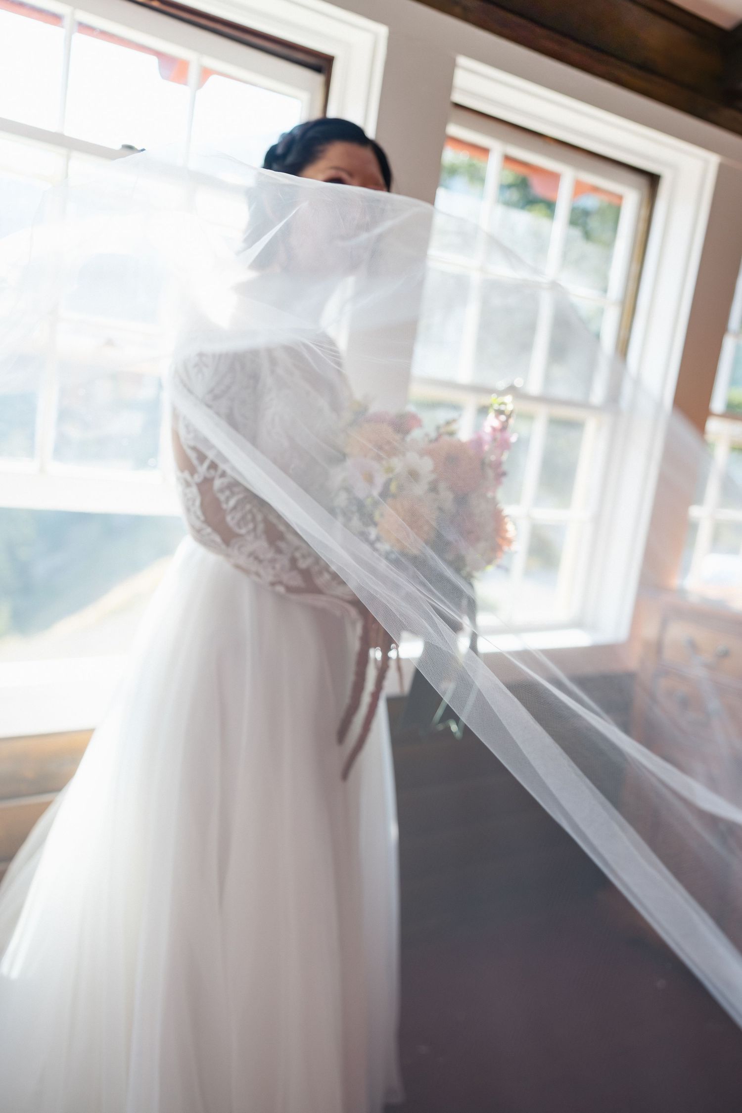 A flowing white wedding dress with delicate lace details hangs by a window in soft natural light.