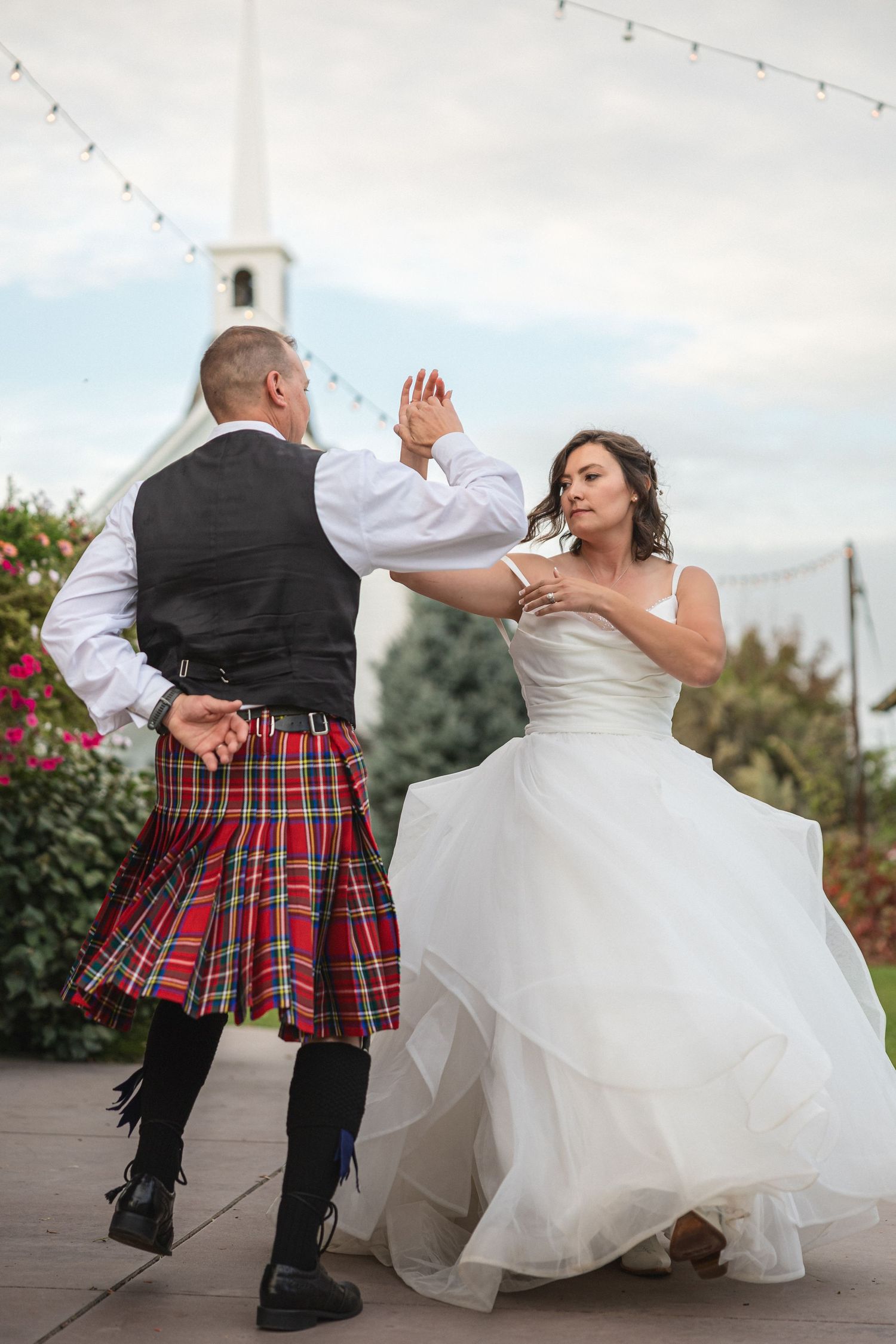 A couple performs a lively dance move on a patio with a white chapel in the background.