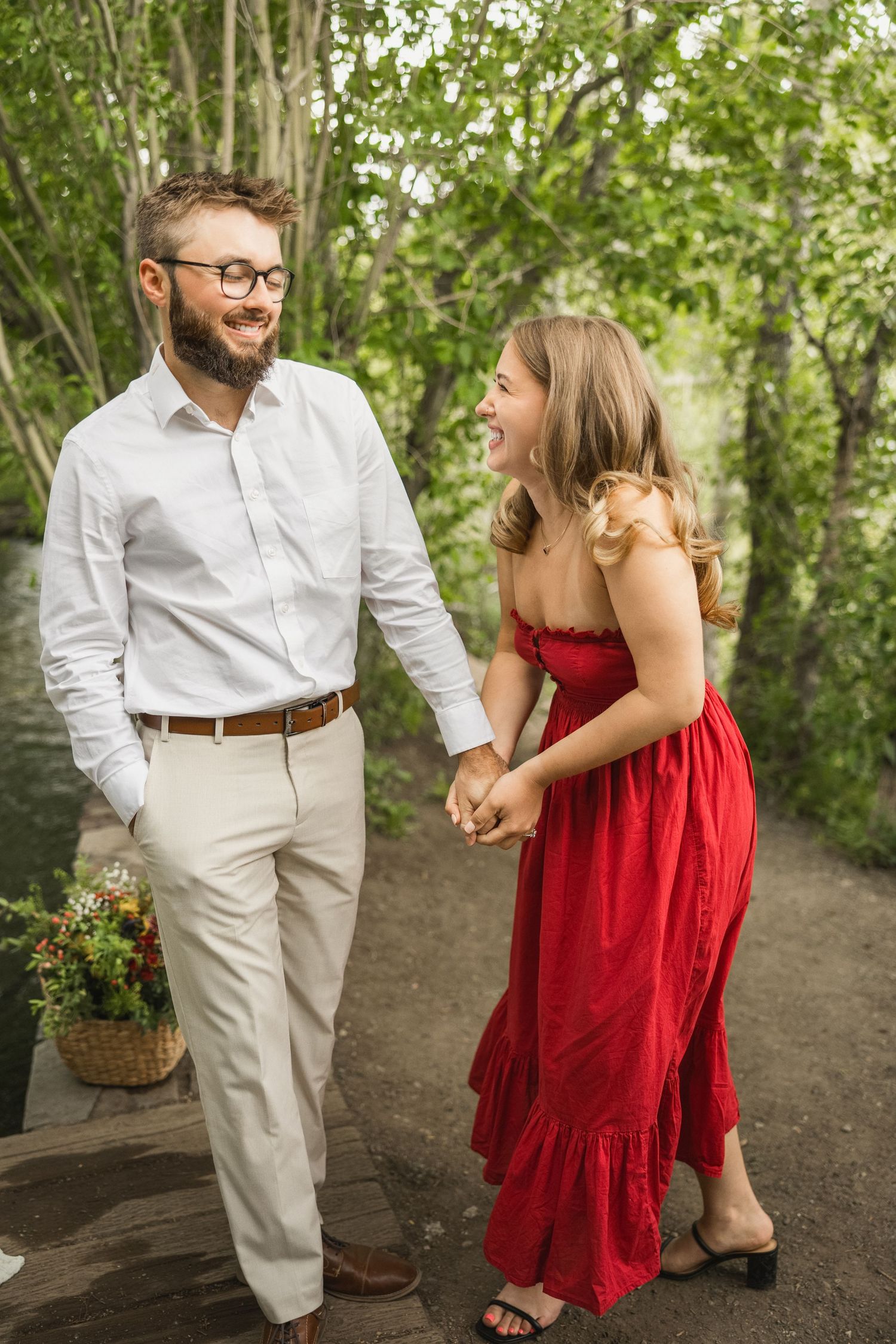 A couple shares a joyful moment on a path outdoors, wearing a white button-down shirt and flowing red dress.