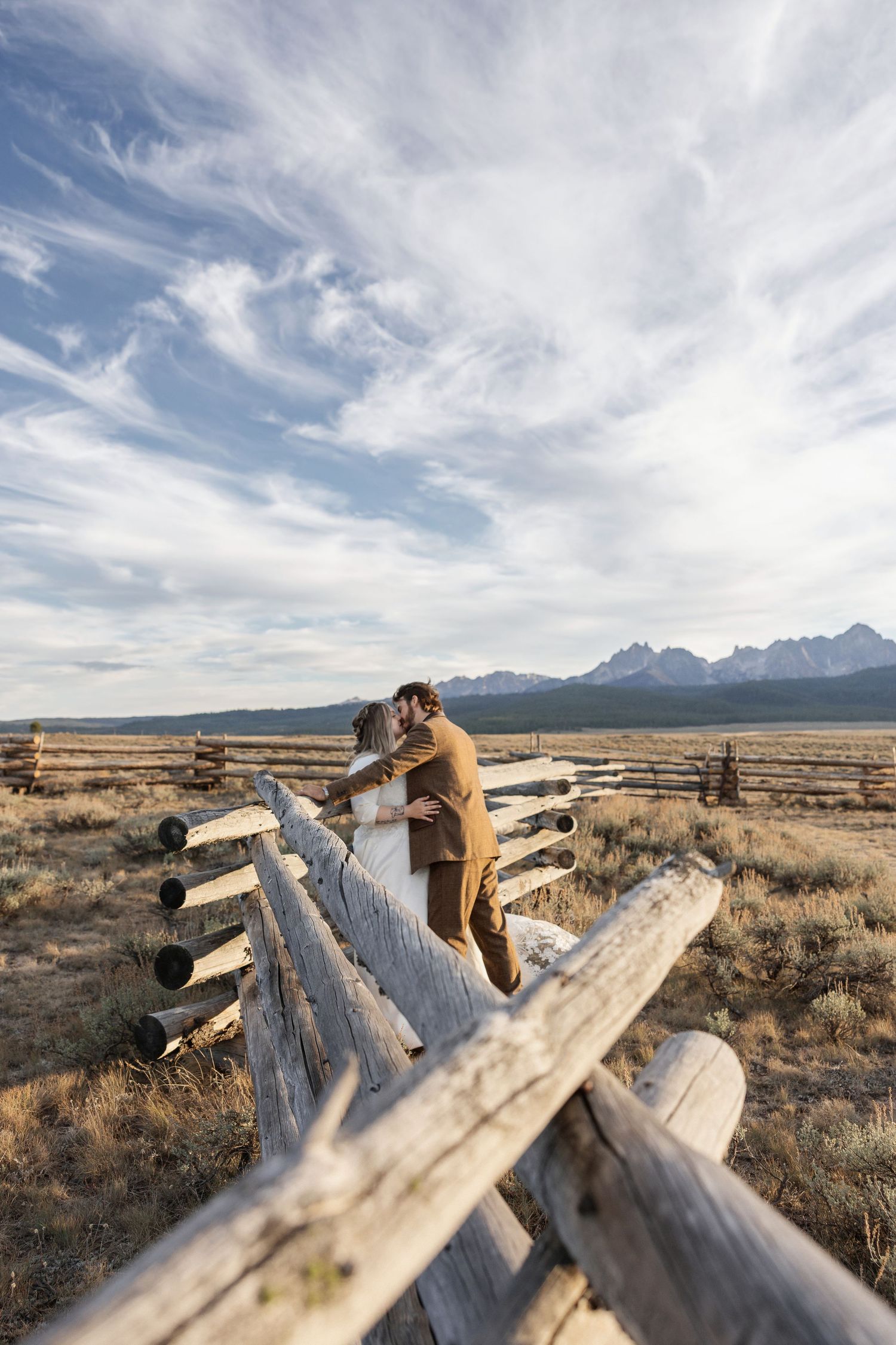 A couple sits together on rustic wooden logs against a dramatic mountain landscape and desert terrain.