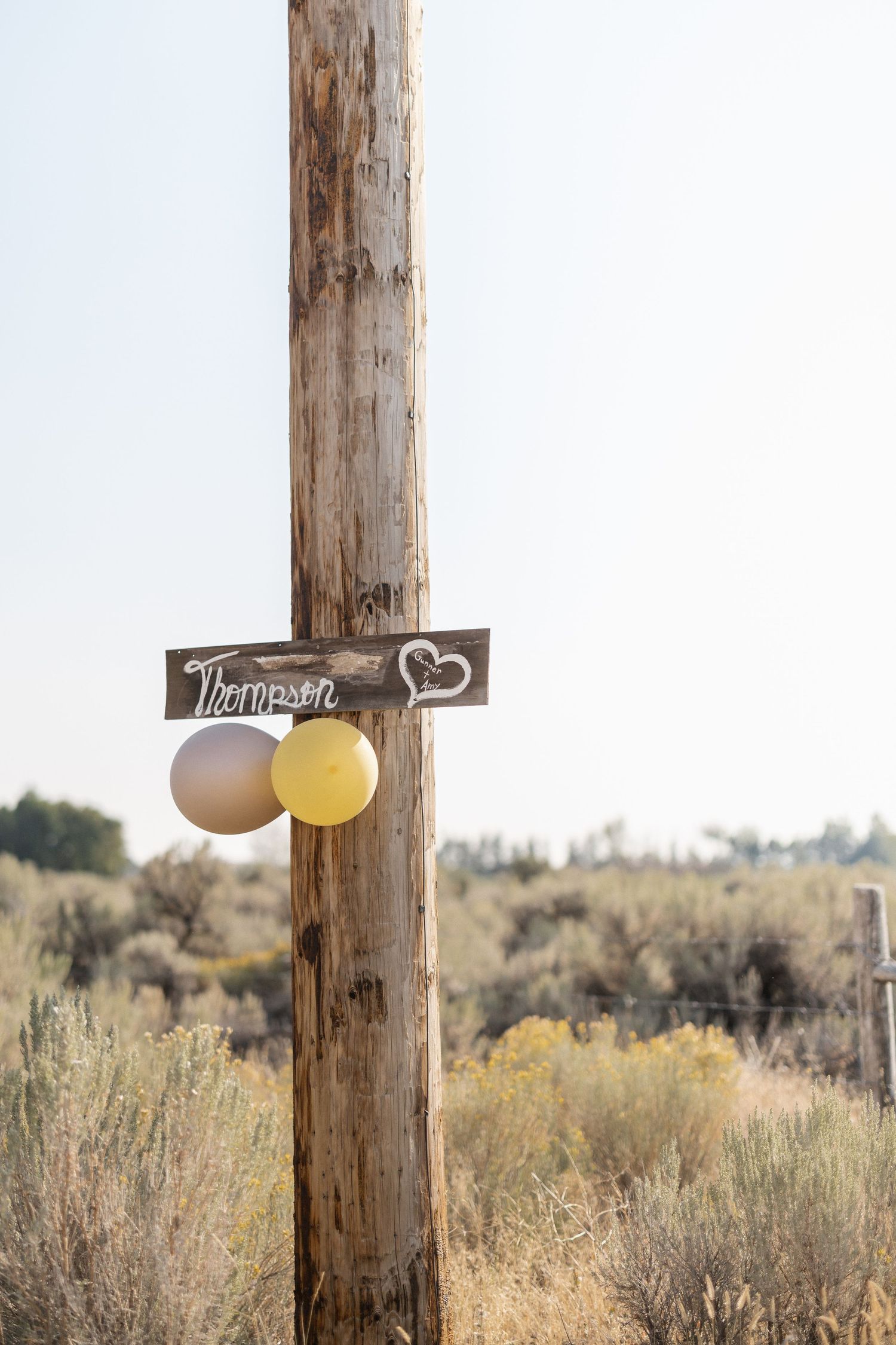 Wooden cross decorated with balloons stands in a desert landscape with sagebrush and distant mountains.