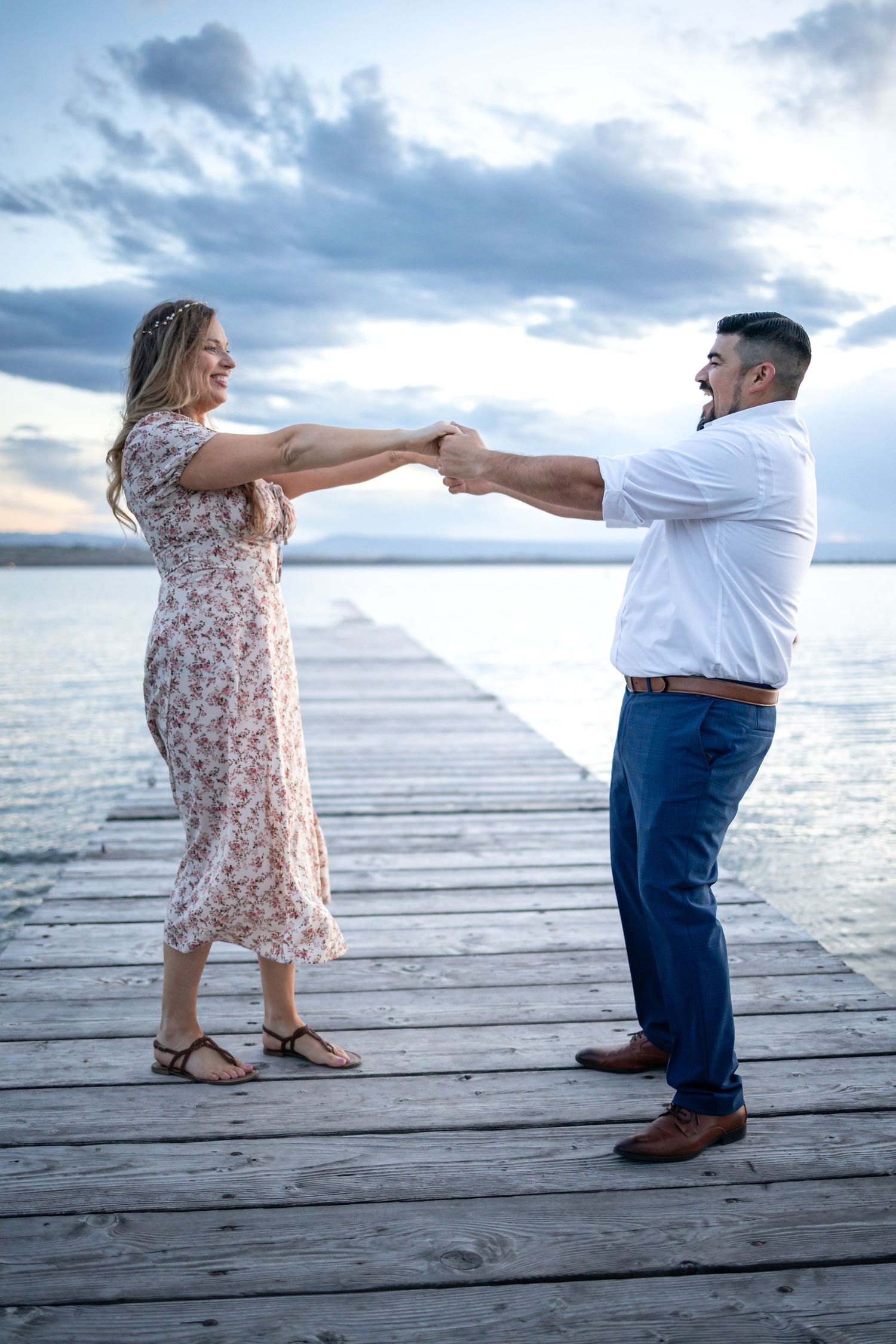 Two people hold hands while dancing playfully on a lakeside dock at dusk with cloudy skies.