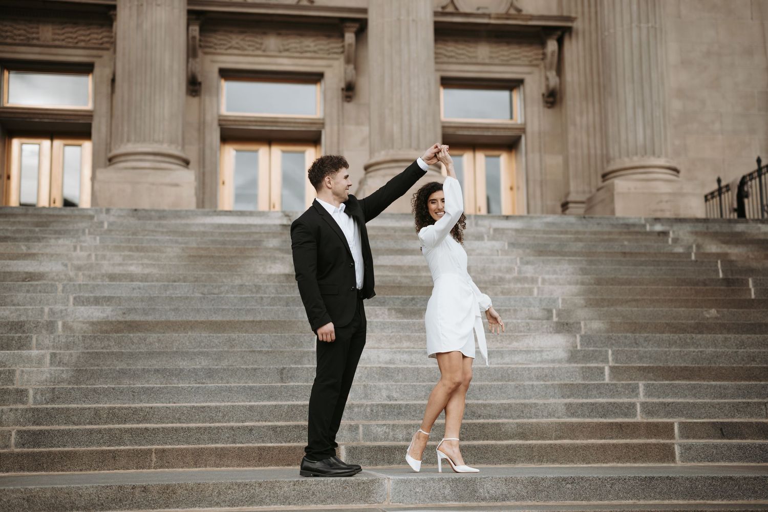 A couple poses playfully on grand stone steps of a classical building.