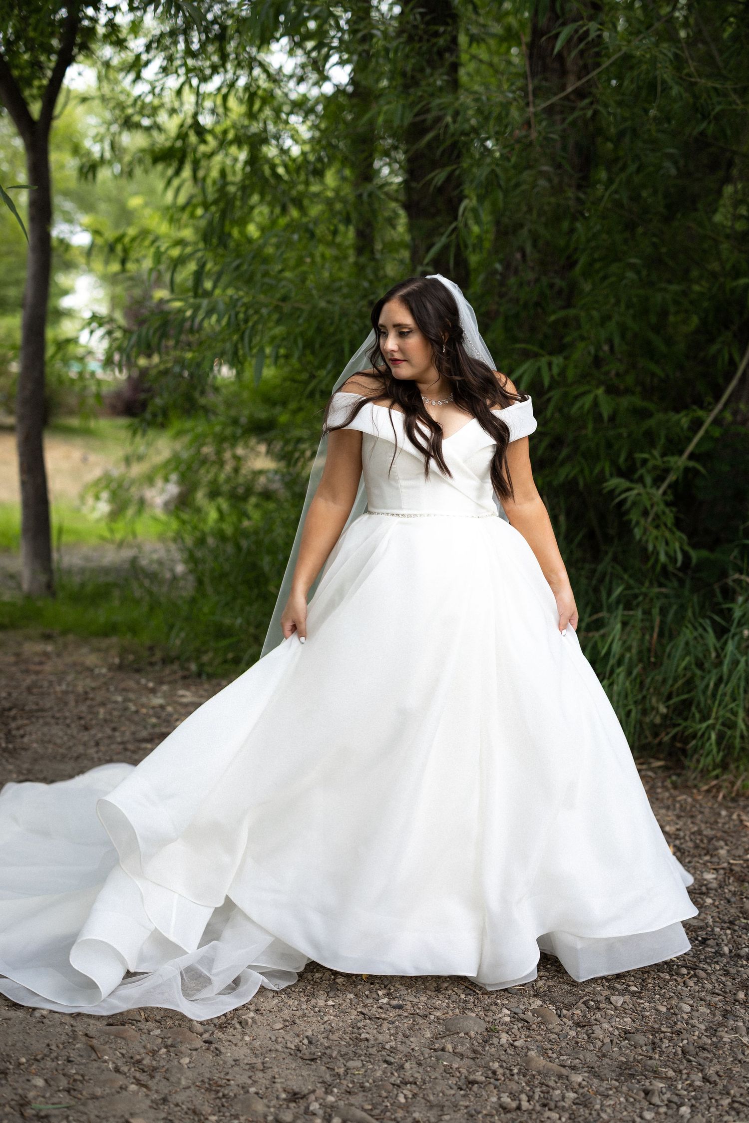 A flowing white wedding gown with tulle fabric spreads across a dirt path in a wooded outdoor setting.