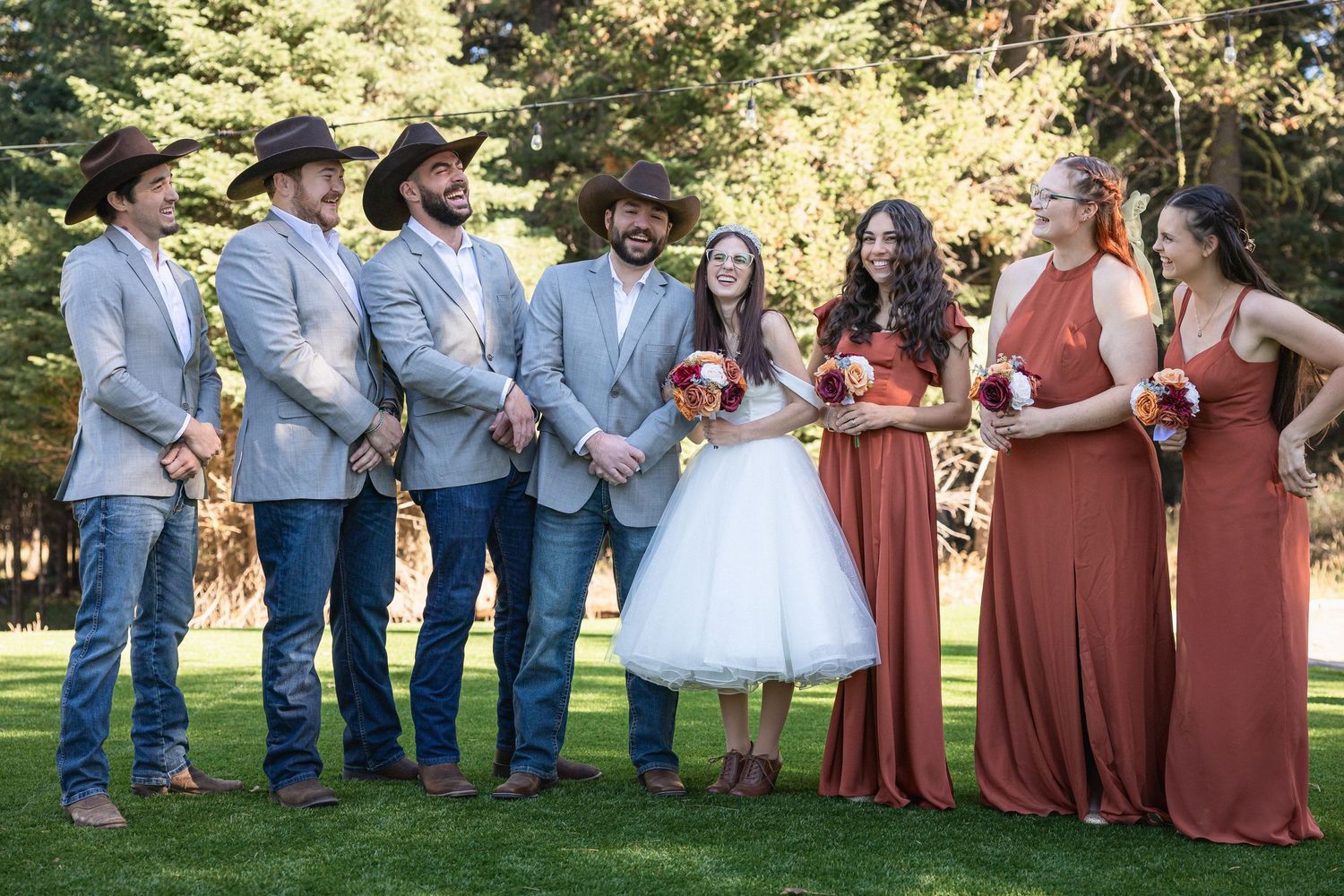 Wedding party stands on grass with groomsmen wearing matching blue suits and black cowboy hats.