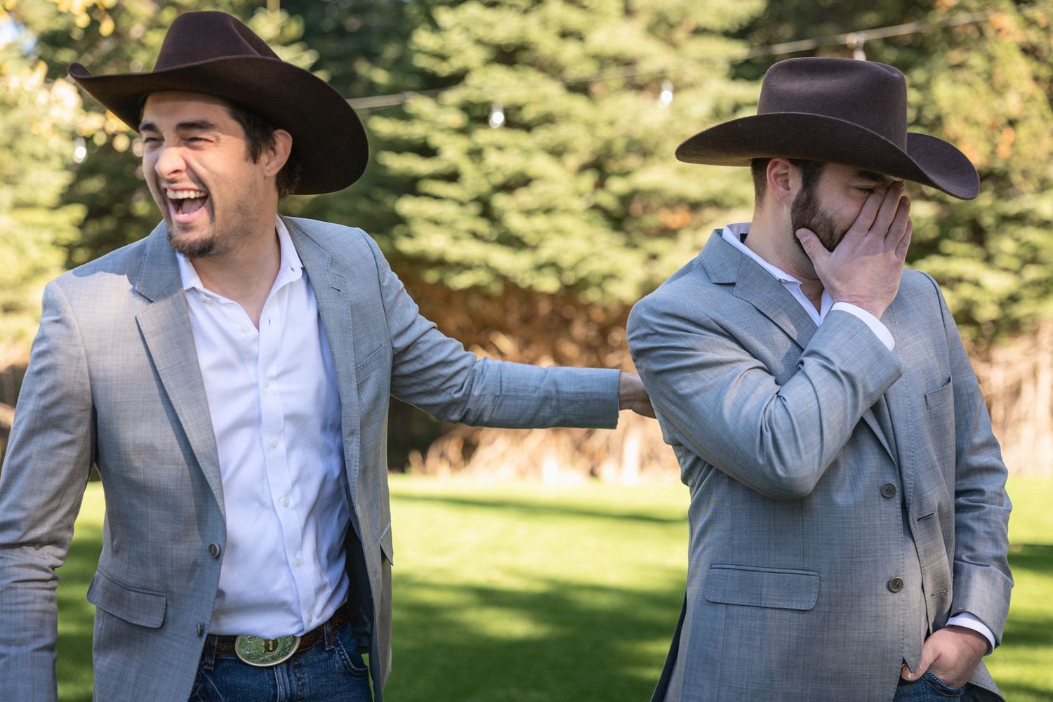 Groomsmen in cowboy hats and gray suits sharing a laugh at outdoor wedding.