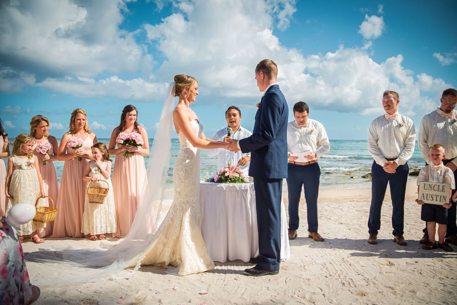 Beachside wedding ceremony takes place on white sand with turquoise ocean and puffy clouds in background.