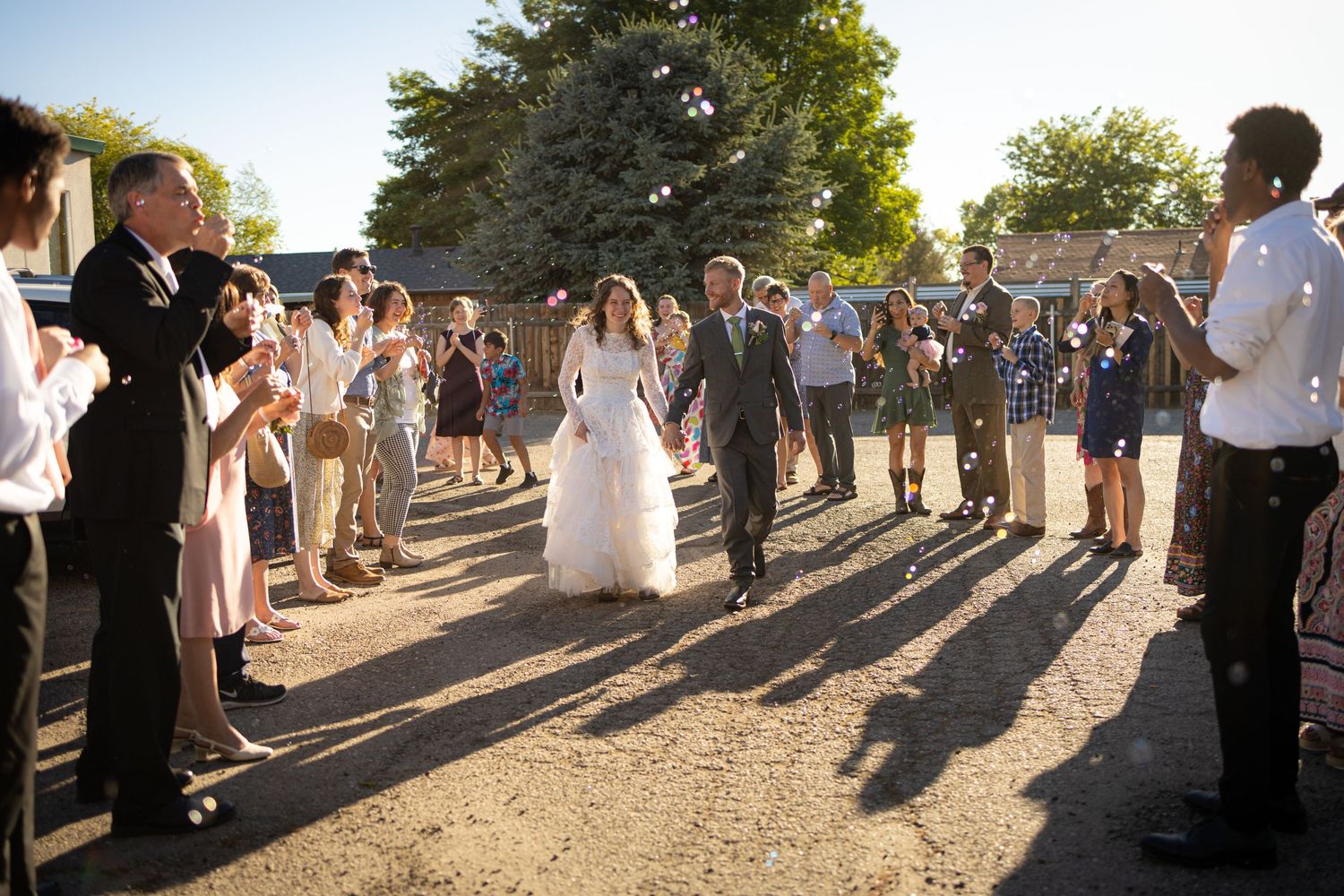 Wedding guests form an aisle outdoors at sunset, creating long shadows while applauding.