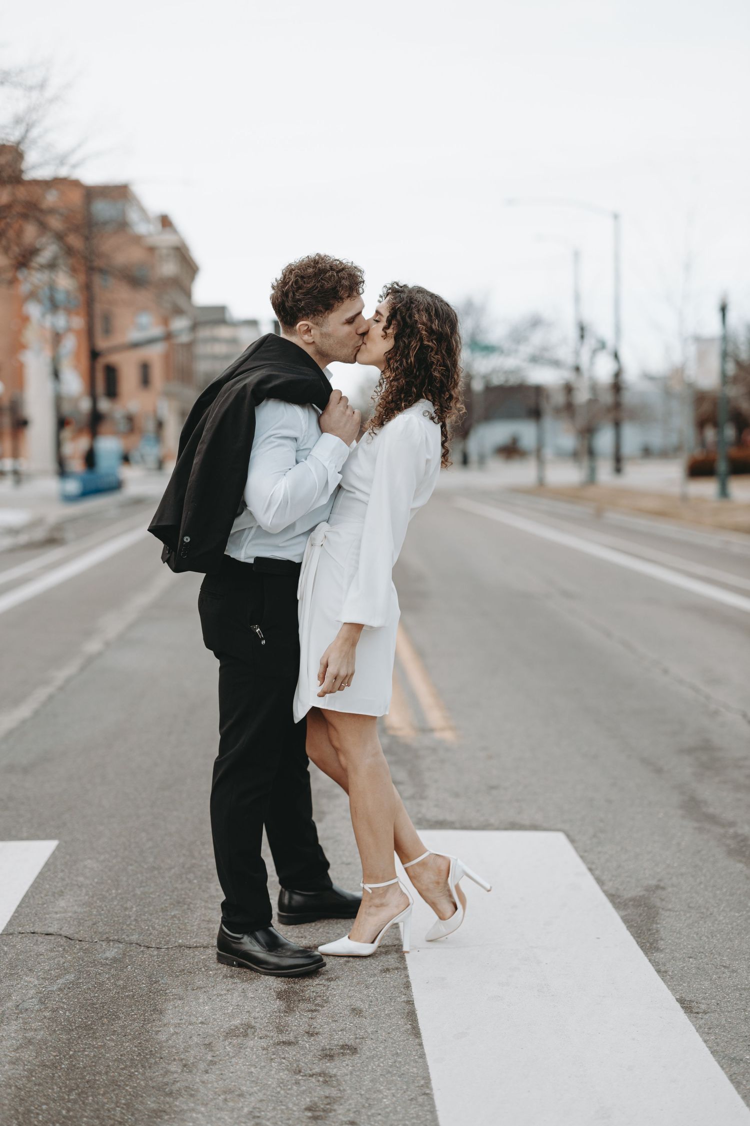 A romantic couple embraces on a crosswalk in an urban setting with brick buildings in background.