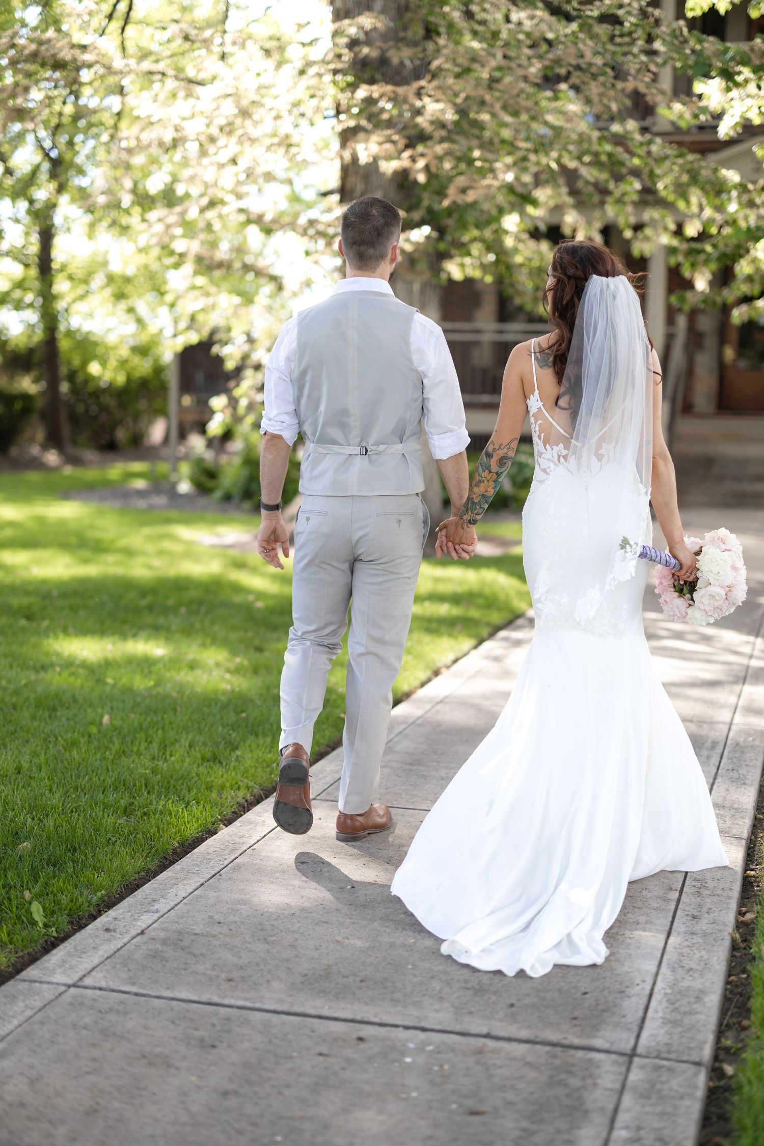 Wedding couple walking away on a concrete path surrounded by green lawn and trees.